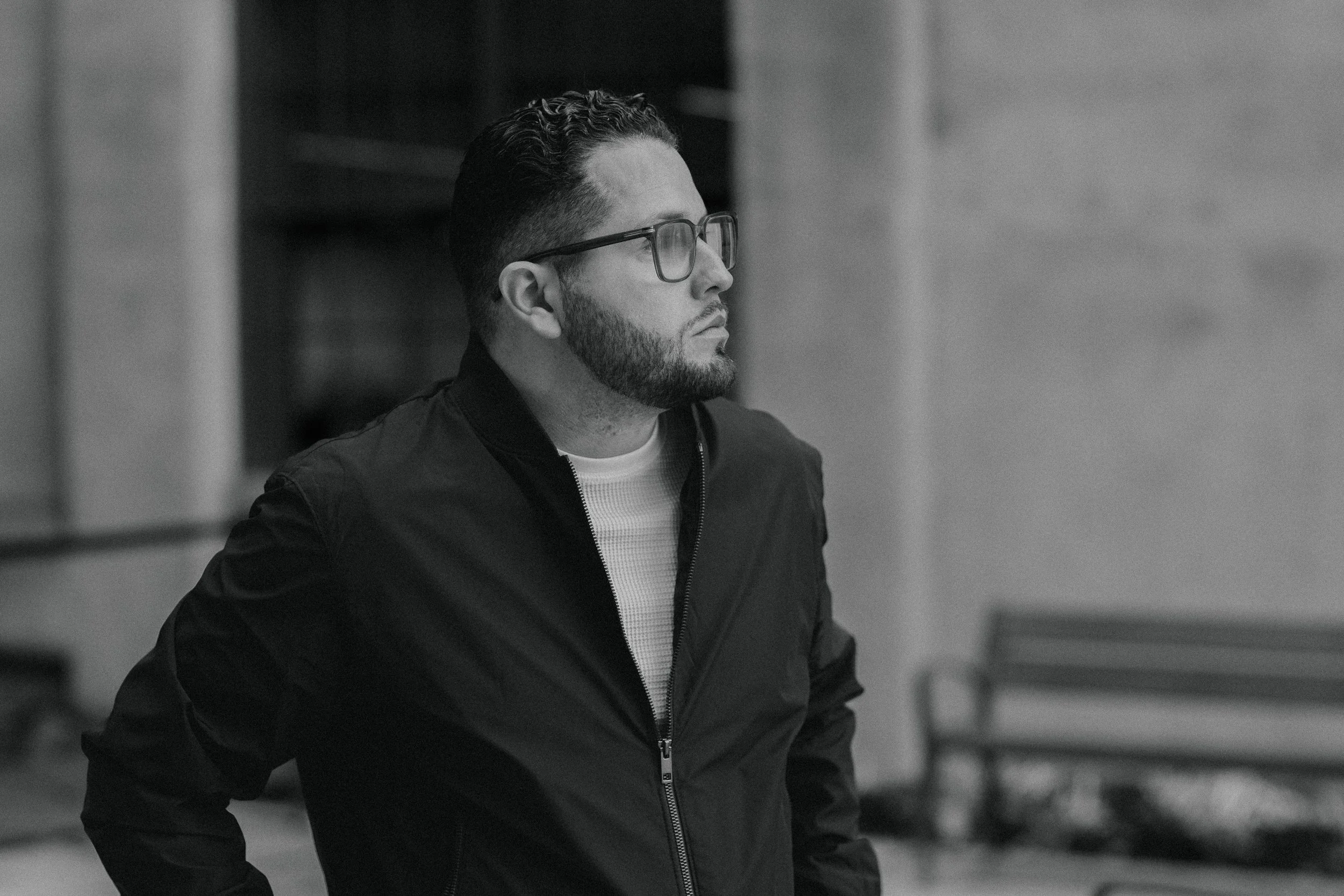 A black and white photo of a man with glasses, a beard, and curly hair, wearing a zip-up jacket, looking to the left.
