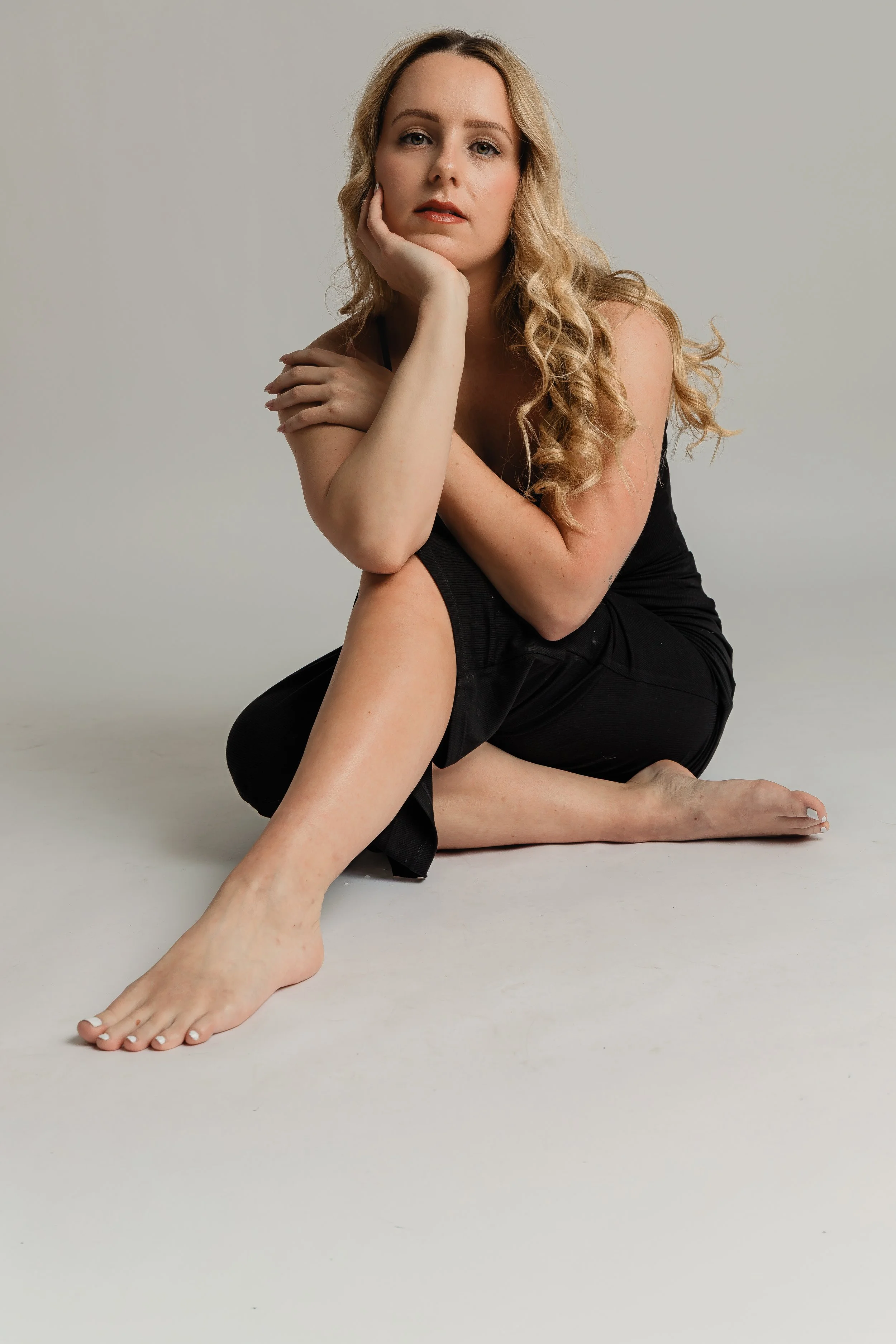 A woman sitting on the ground with her legs crossed, resting her head on her hand, wearing a black sleeveless dress, with long wavy blonde hair, in front of a plain light-colored background.