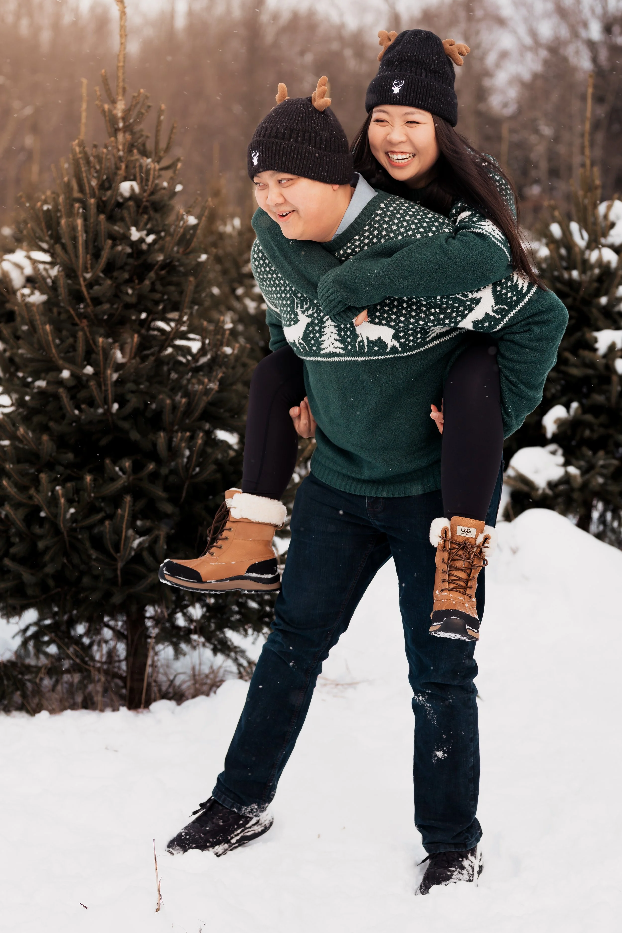 A smiling couple in winter clothes, with the woman on the man’s back, standing in a snowy forest with evergreen trees.