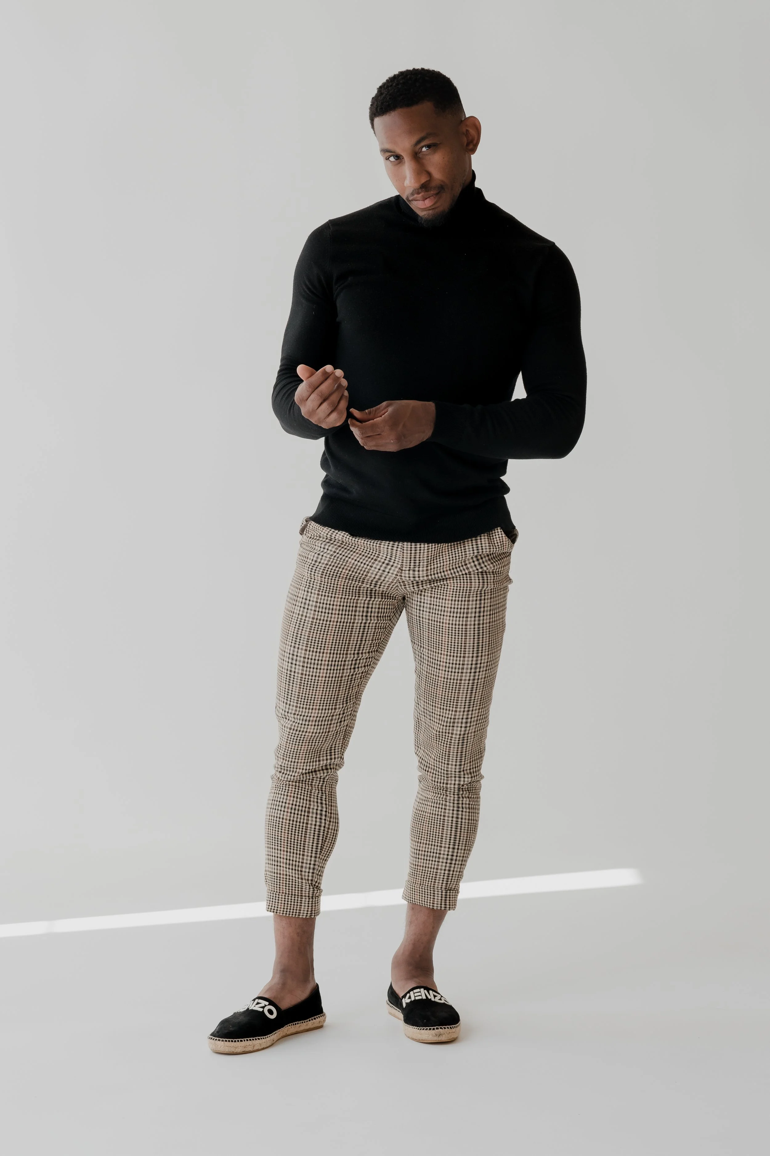 A man stands against a plain light grey wall, dressed in a fitted black turtleneck, checkered pants, and black espadrille shoes with white lettering, looking confidently at the camera.