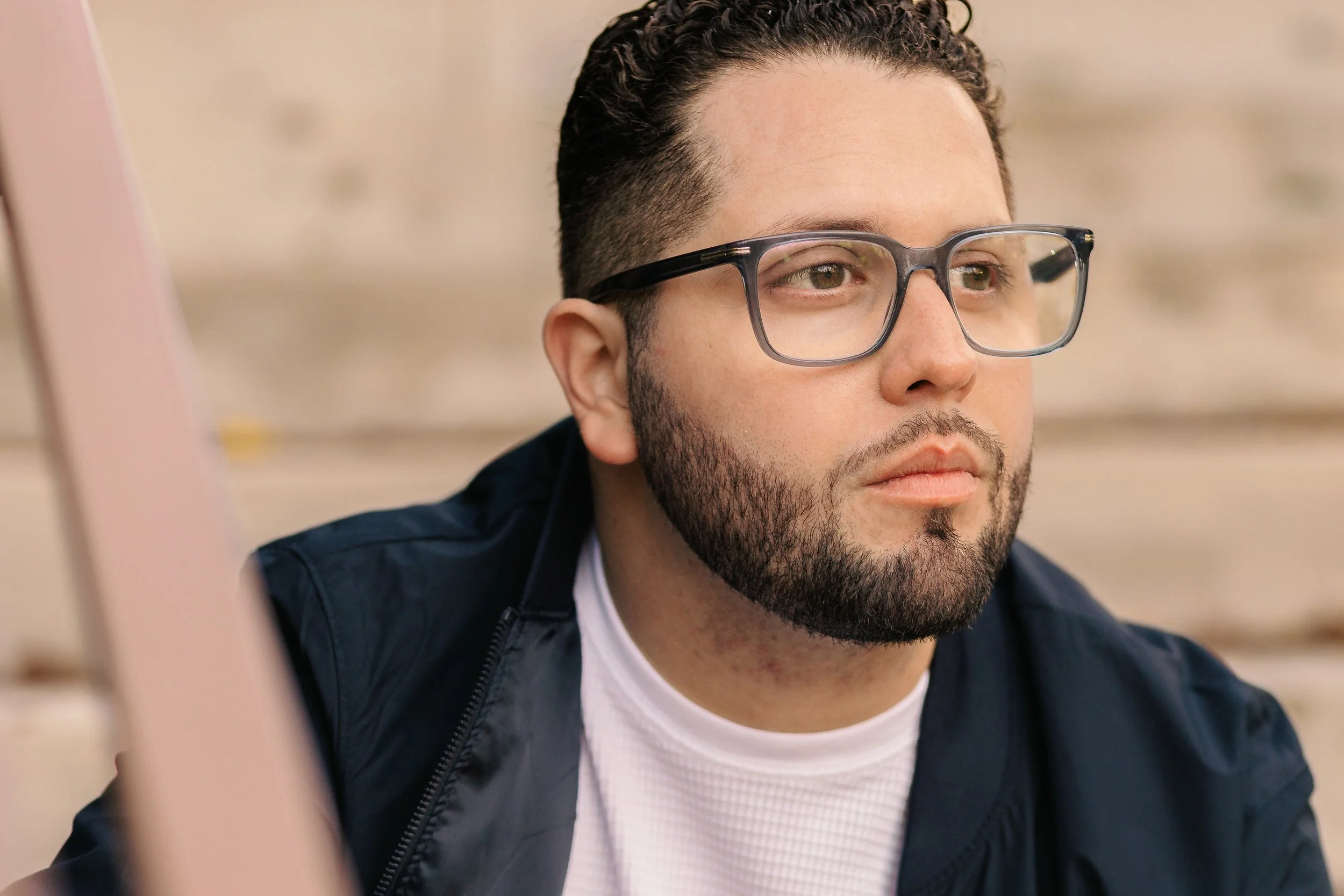 A man with glasses and a beard looking pensively to the side, sitting outdoors near a beige wall.