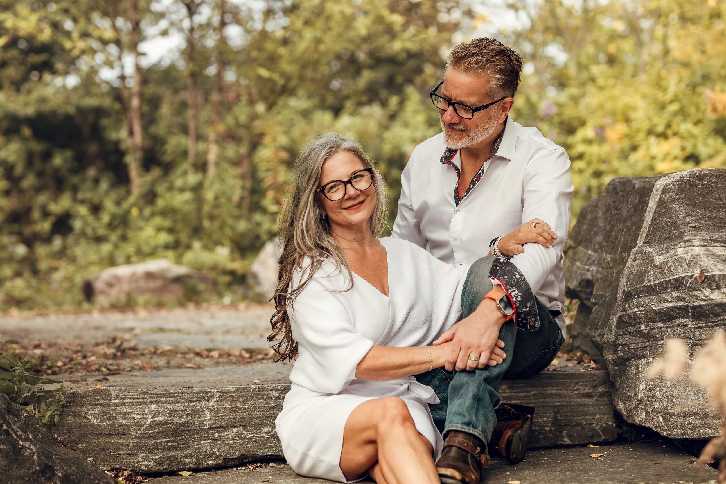 A smiling middle-aged couple with gray hair and glasses sitting outdoors on a wooden log, surrounded by trees and rocks, during autumn.
