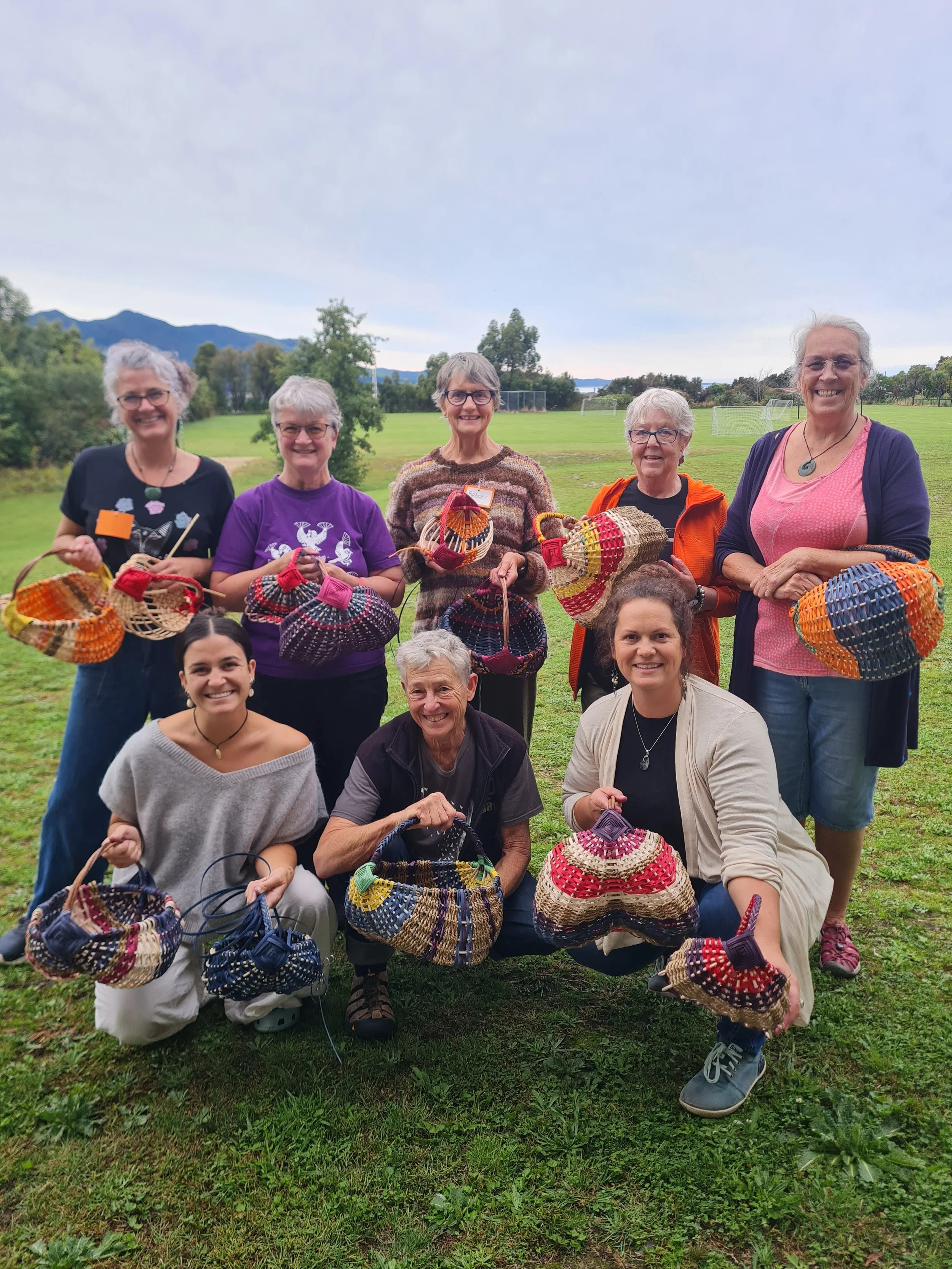Group of women and one man outdoors on a grassy field, holding colorful woven baskets, with trees and mountains in the background.