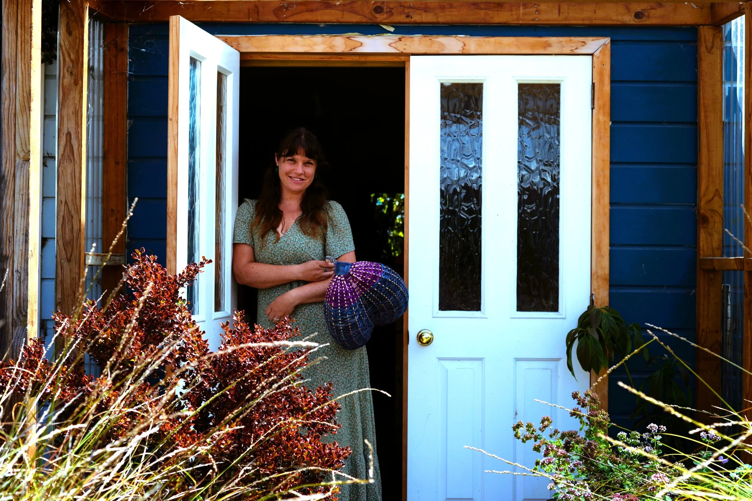 A woman standing in a doorway of a house, holding a colorful woven basket, surrounded by plants and greenery.