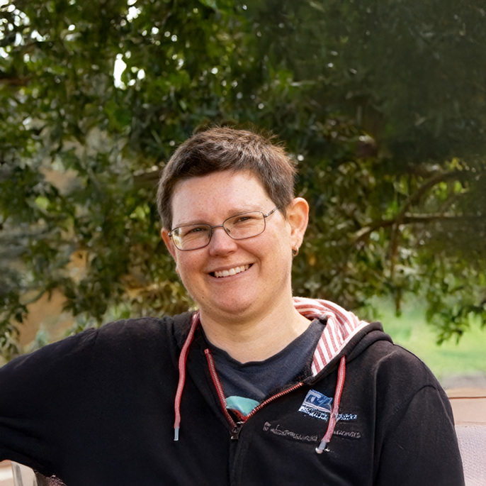 A woman with short dark hair and glasses smiling outdoors in front of a tree.