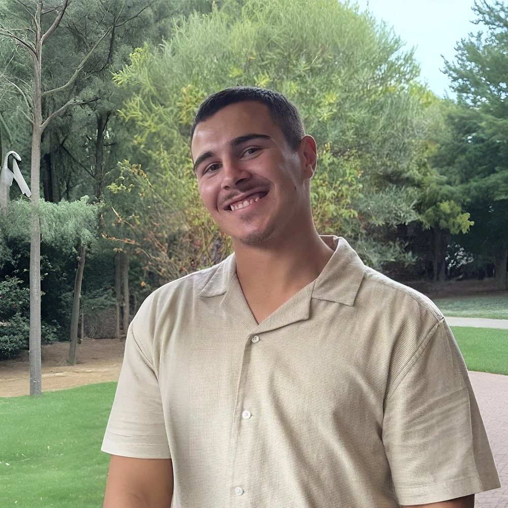 A young man with short dark hair, wearing a beige short-sleeved collared shirt, smiling outdoors in a park with trees, grass, and a pathway in the background.