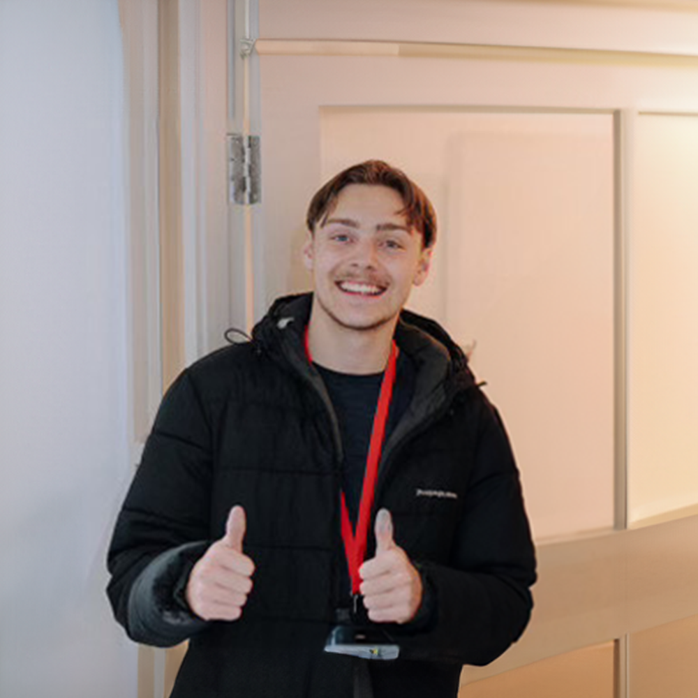 Young man smiling and giving two thumbs up, wearing a black jacket and red lanyard, standing indoors near a door.