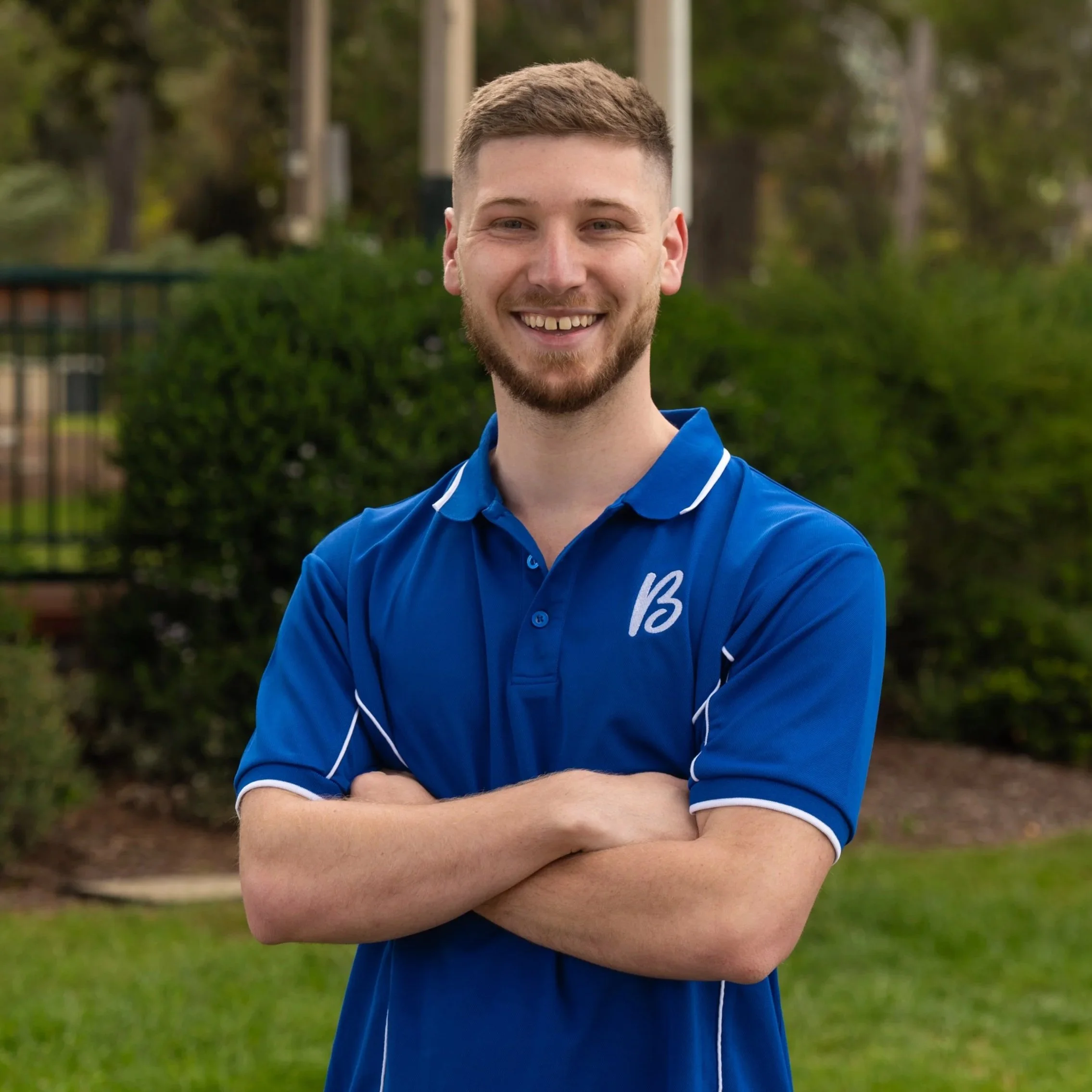 A young man with short brown hair, a beard, and a smile, wearing a blue hoodie with a white logo on the chest, stands in front of a leafy green bush outdoor during daytime.