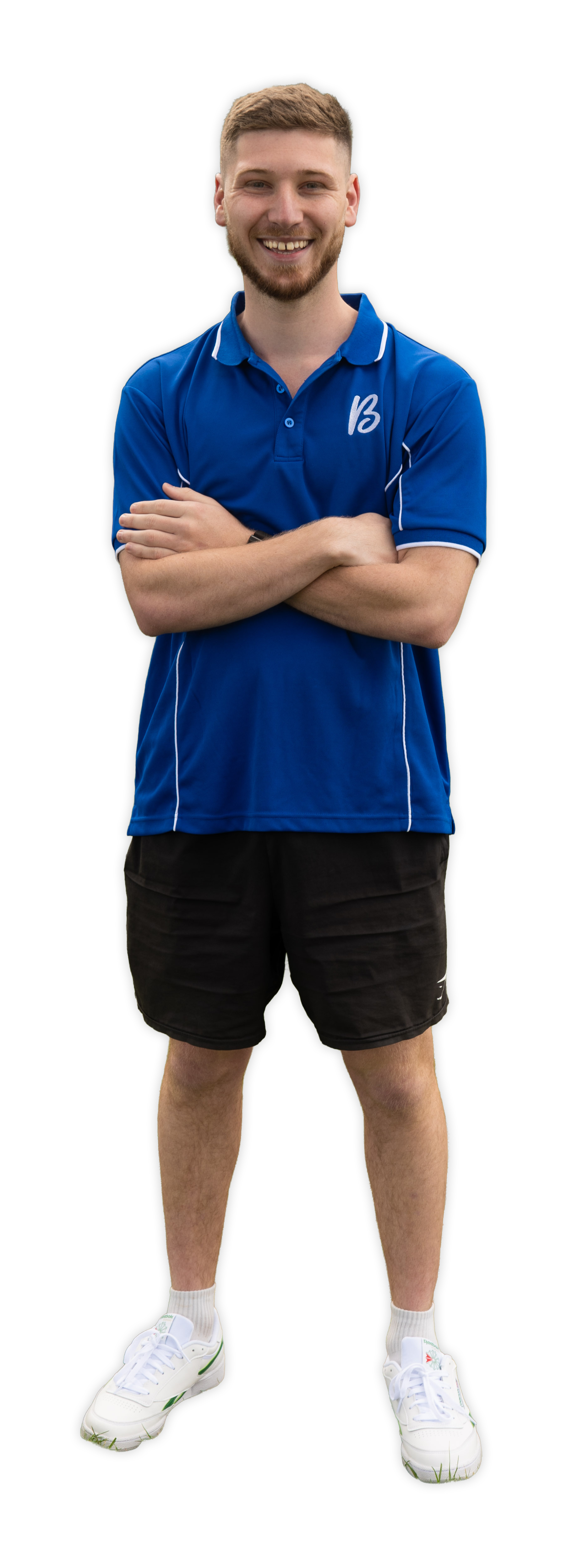 A young man with short light brown hair and a beard, smiling with arms crossed, wearing a blue sports shirt, black shorts, white sneakers, and white socks.