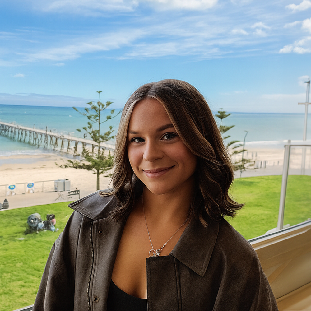 A woman smiling indoors with a view of a beach and pier outside the window behind her.