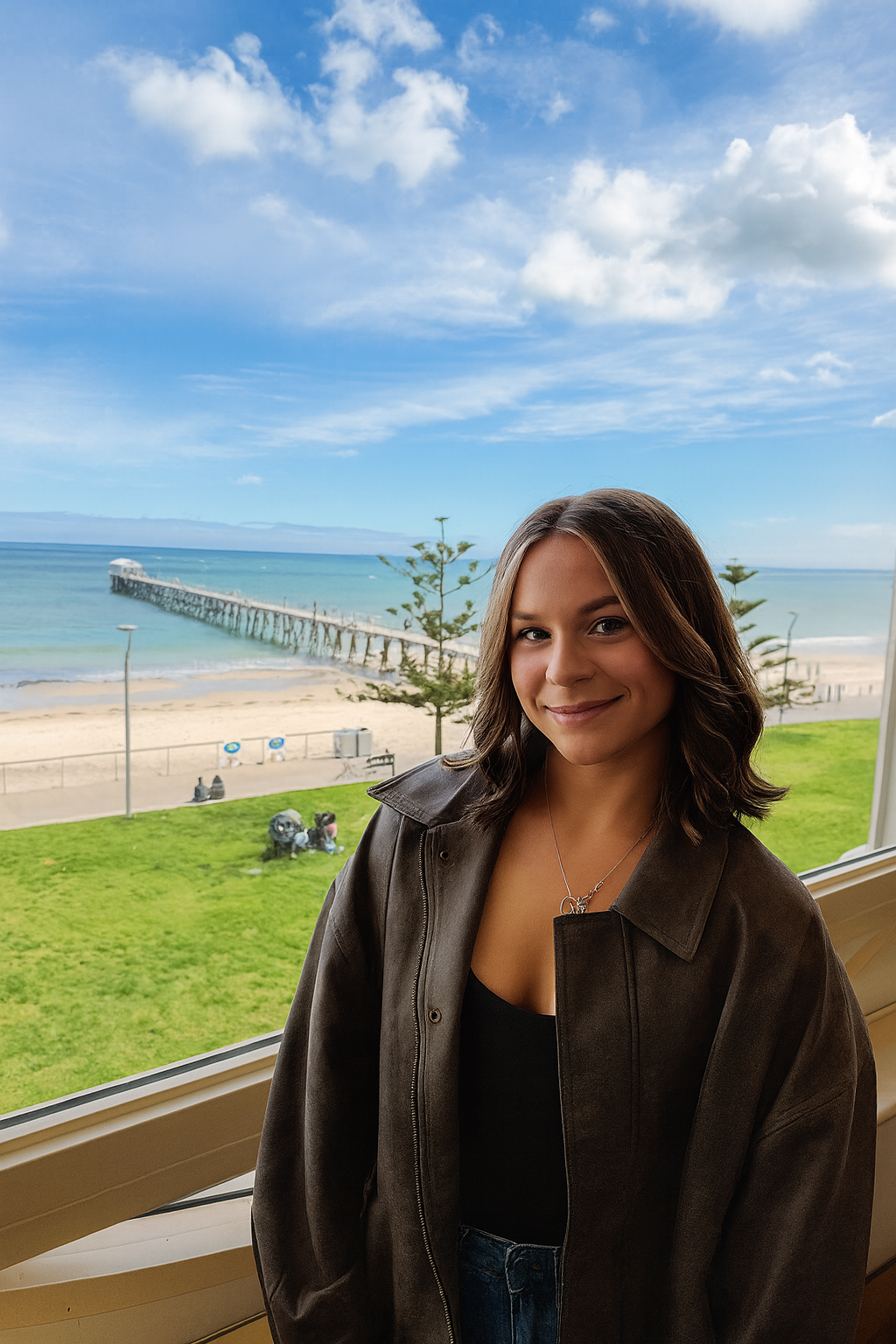 Young woman smiling indoors with large window overlooking a beach with a pier, sandy shore, and blue sky with clouds.