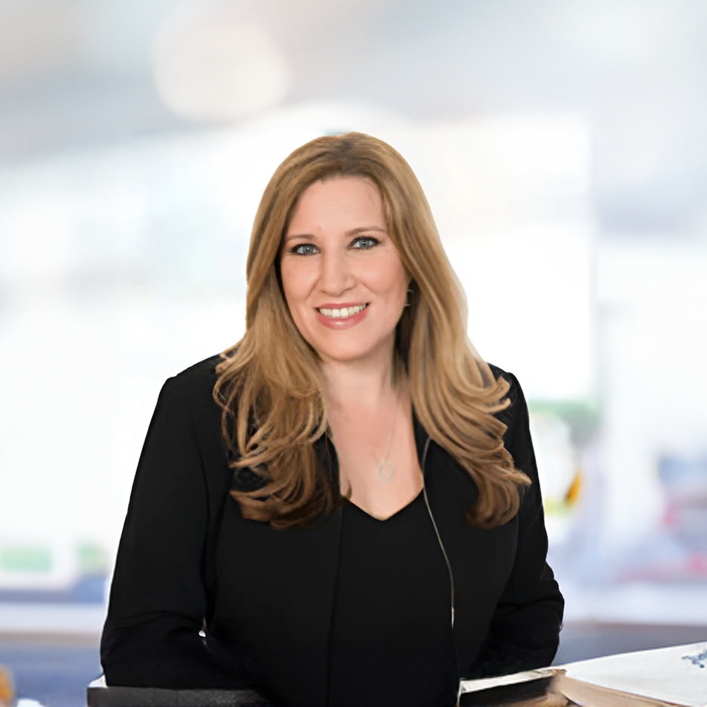 A woman with long blonde hair, wearing a black blazer, smiling and sitting at a desk in a bright office setting.