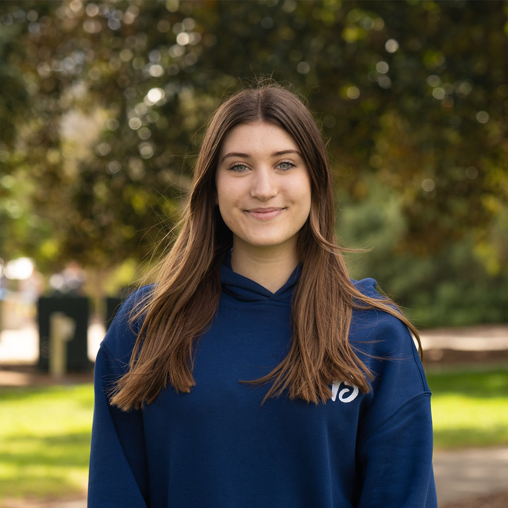 Young woman with long brown hair wearing a blue hoodie, standing outdoors with a blurred background of trees and grass.