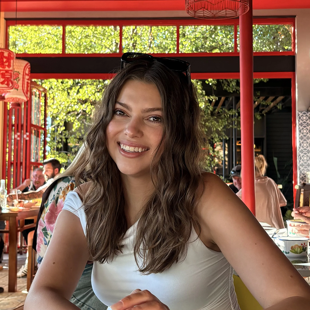 Young woman with long wavy brown hair smiling at a restaurant table, wearing a white sleeveless top, with sunglasses on her head, in a decorated Asian-themed restaurant with red framing, hanging lanterns, and other patrons in the background.