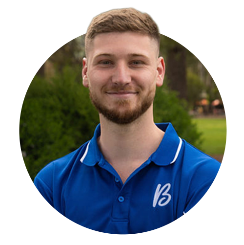A young man with light brown hair and a beard smiling, wearing a blue polo shirt with a logo, outdoors in a park with trees and grass.