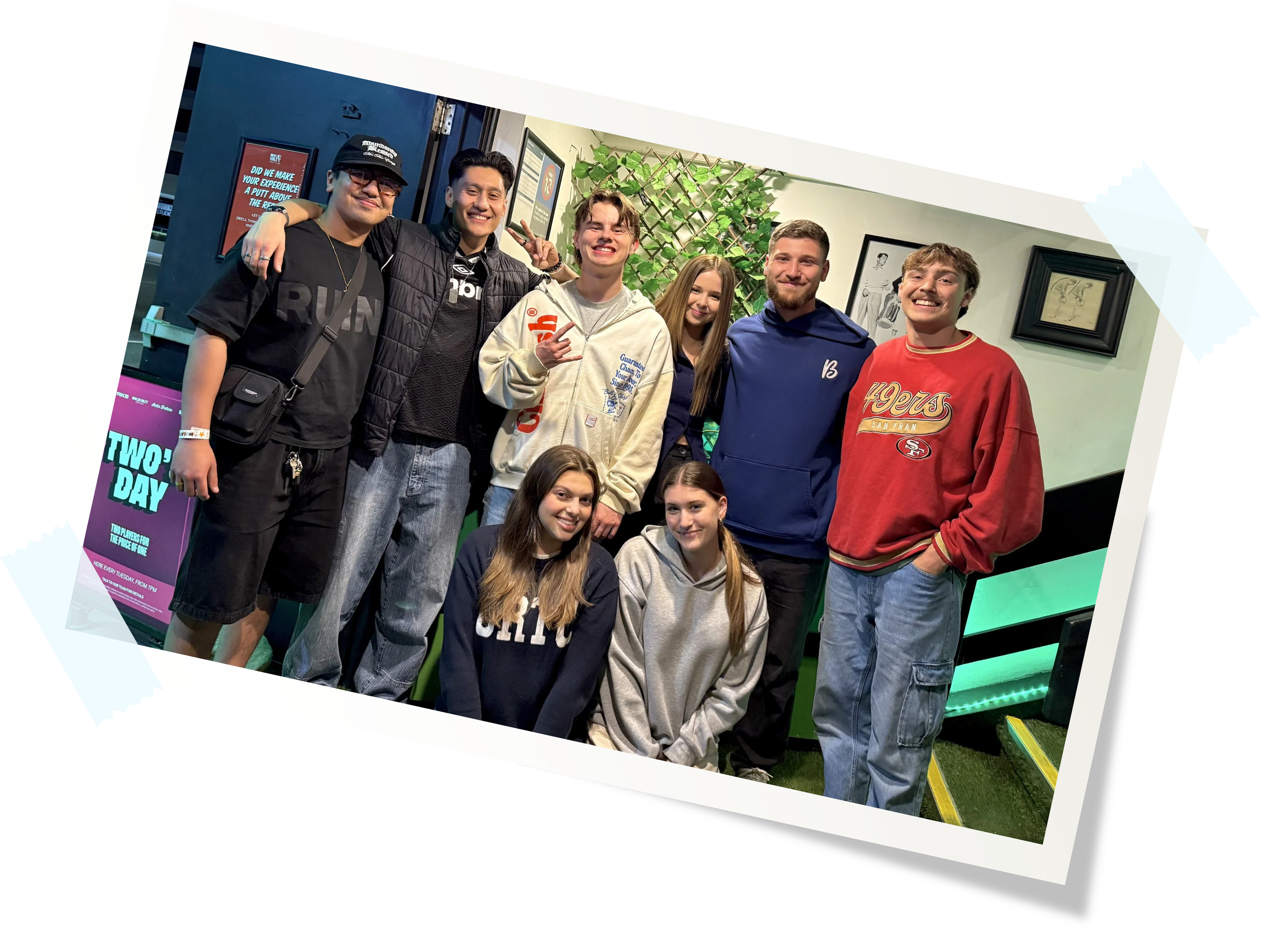 Group of nine young people posing indoors, some standing and some kneeling, smiling at the camera.