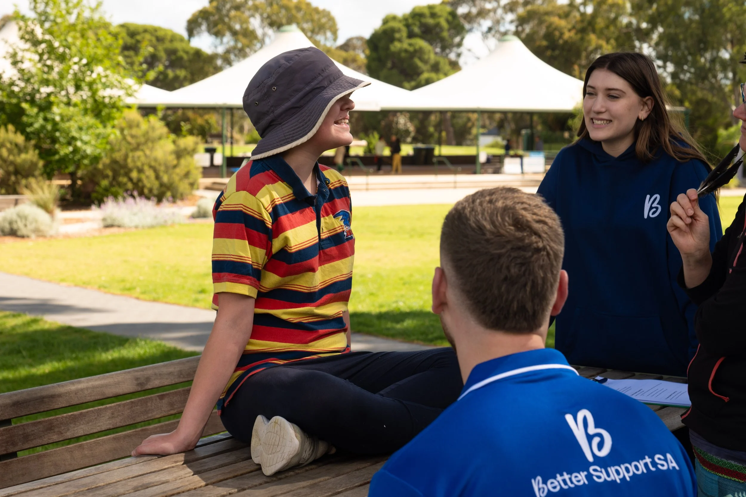 Group of young people in a park, talking and smiling near a wooden bench. Two are standing, one sitting on the bench with legs crossed, and one with their back to the camera. The park has green grass, trees, and white tents in the background.