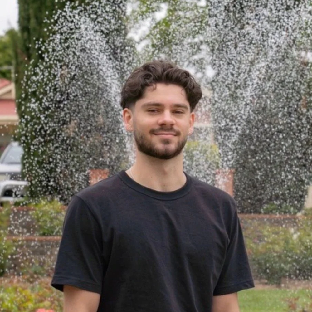 A young man with dark, curly hair and a beard, wearing a black T-shirt, standing outdoors in front of a water fountain with water splashing around him. There are trees and houses in the background.