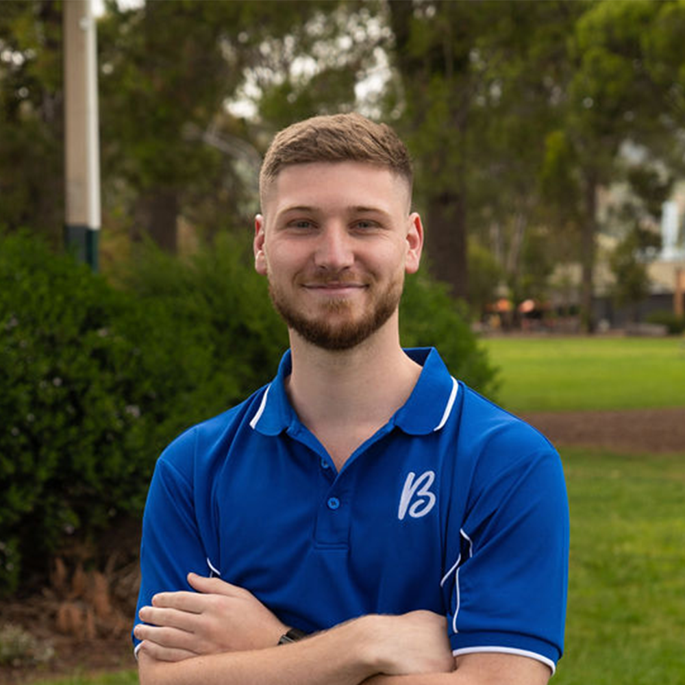 A young man with a beard, wearing a blue polo shirt, smiling with arms crossed in an outdoor park setting.