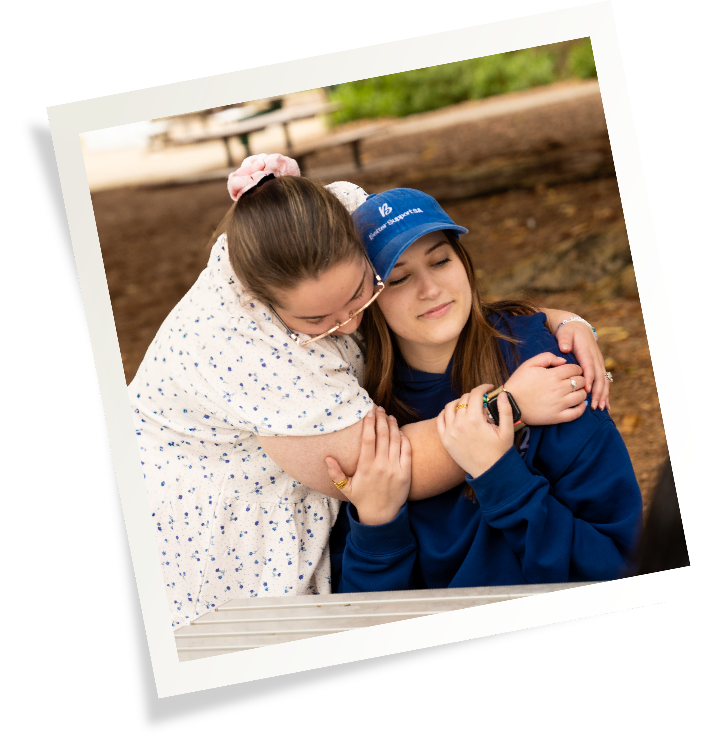 Two women hugging each other, one wearing a blue cap and the other wearing glasses and a white polka dot dress, outdoors with a dirt path and benches in the background.