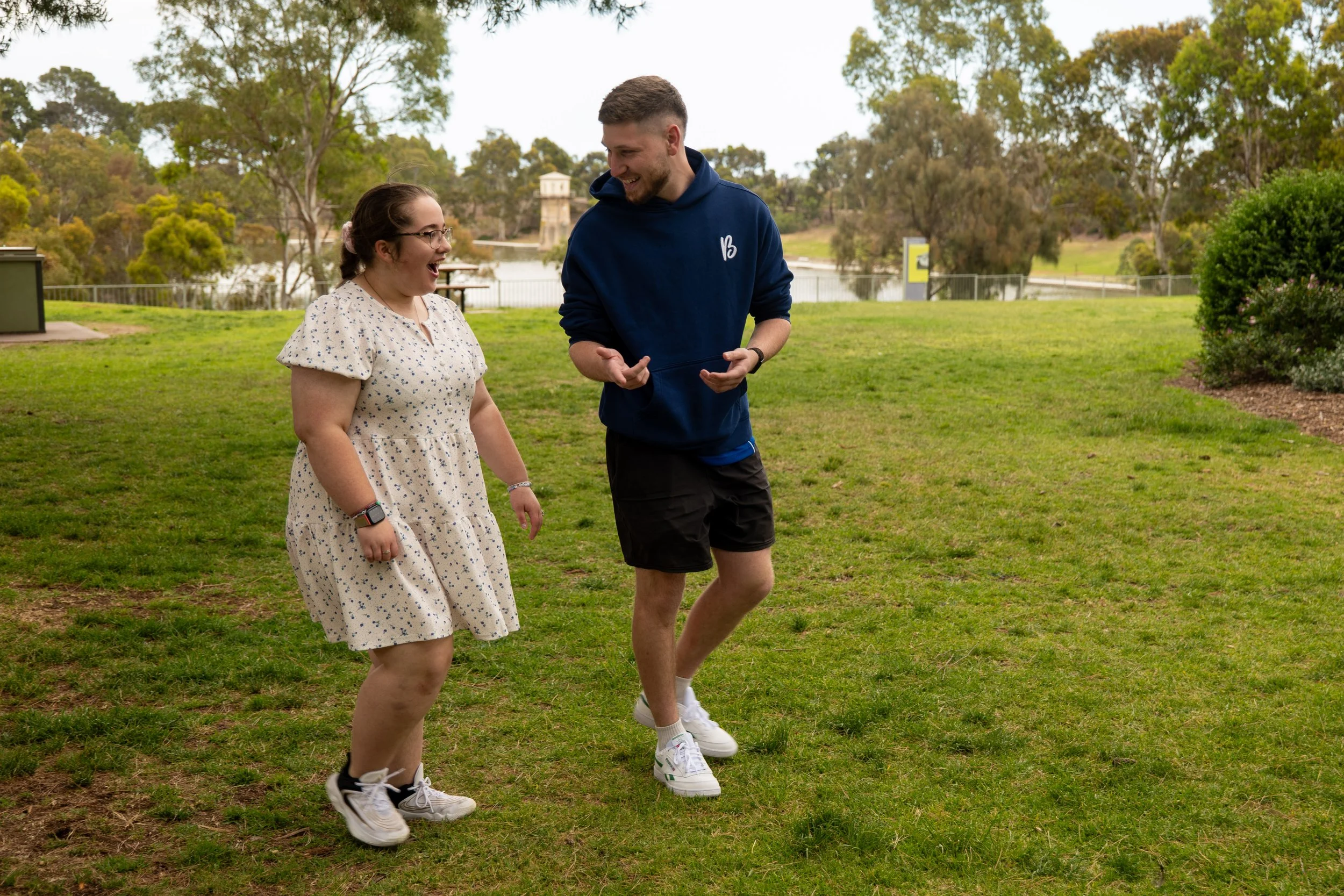 Two people, a woman and a man, are outdoors on a grassy area near a lake, smiling and interacting with each other. The woman has glasses, is wearing a beige dress with small dark patterns, and white sneakers. The man is wearing a dark blue hoodie, dark shorts, and white sneakers. The background features trees, water, and a cloudy sky.