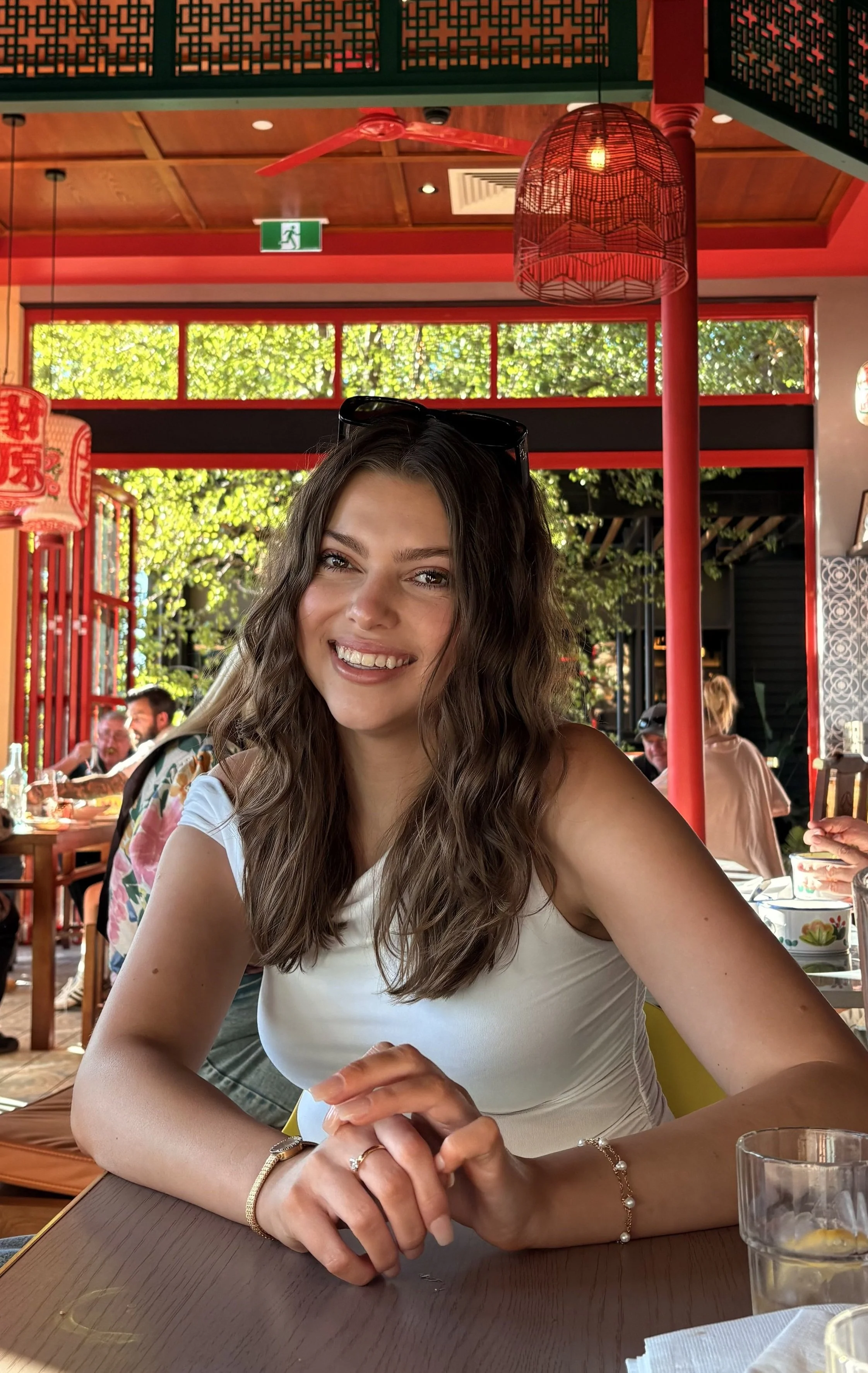 A smiling woman with wavy brown hair wearing a white shirt, sitting at a table in a restaurant with red decor and greenery outside.