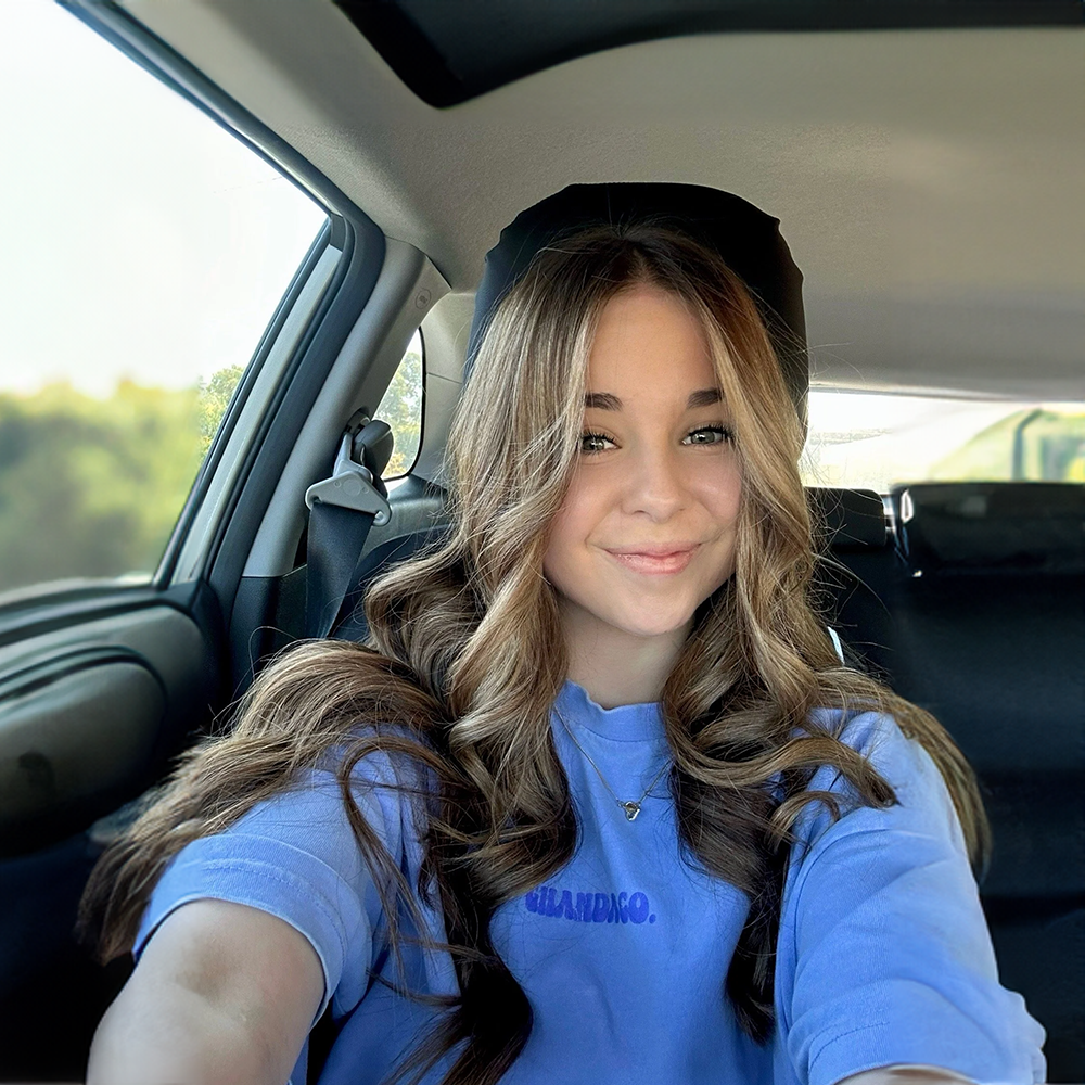 Young woman with long, wavy brown hair, smiling and sitting inside a vehicle, with a scenic outdoors background visible through the window.