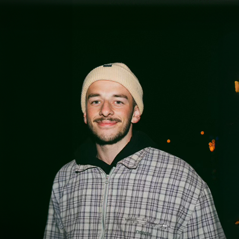 A young man with a beard wearing a beige beanie, black hoodie, and plaid jacket, smiling at the camera against a dark background with faint lights.