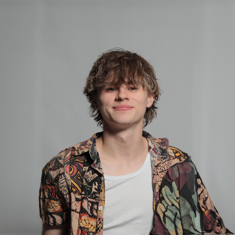 Young man with wavy brown hair wearing a colorful patterned shirt and a white t-shirt, smiling against a plain gray background.