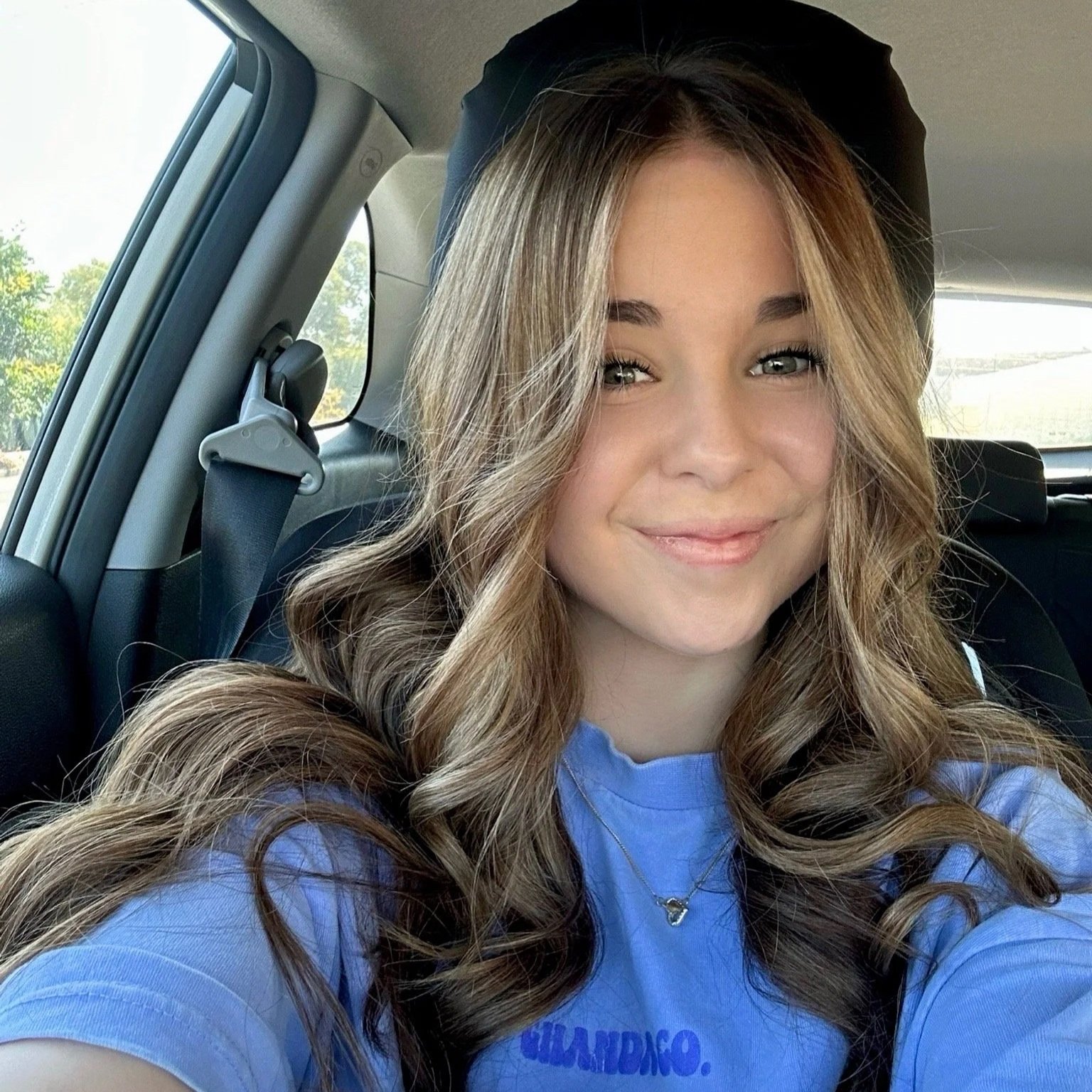 Young woman with long wavy brown hair, smiling, wearing a light blue shirt and a small necklace, sitting in a vehicle with trees visible through the window.