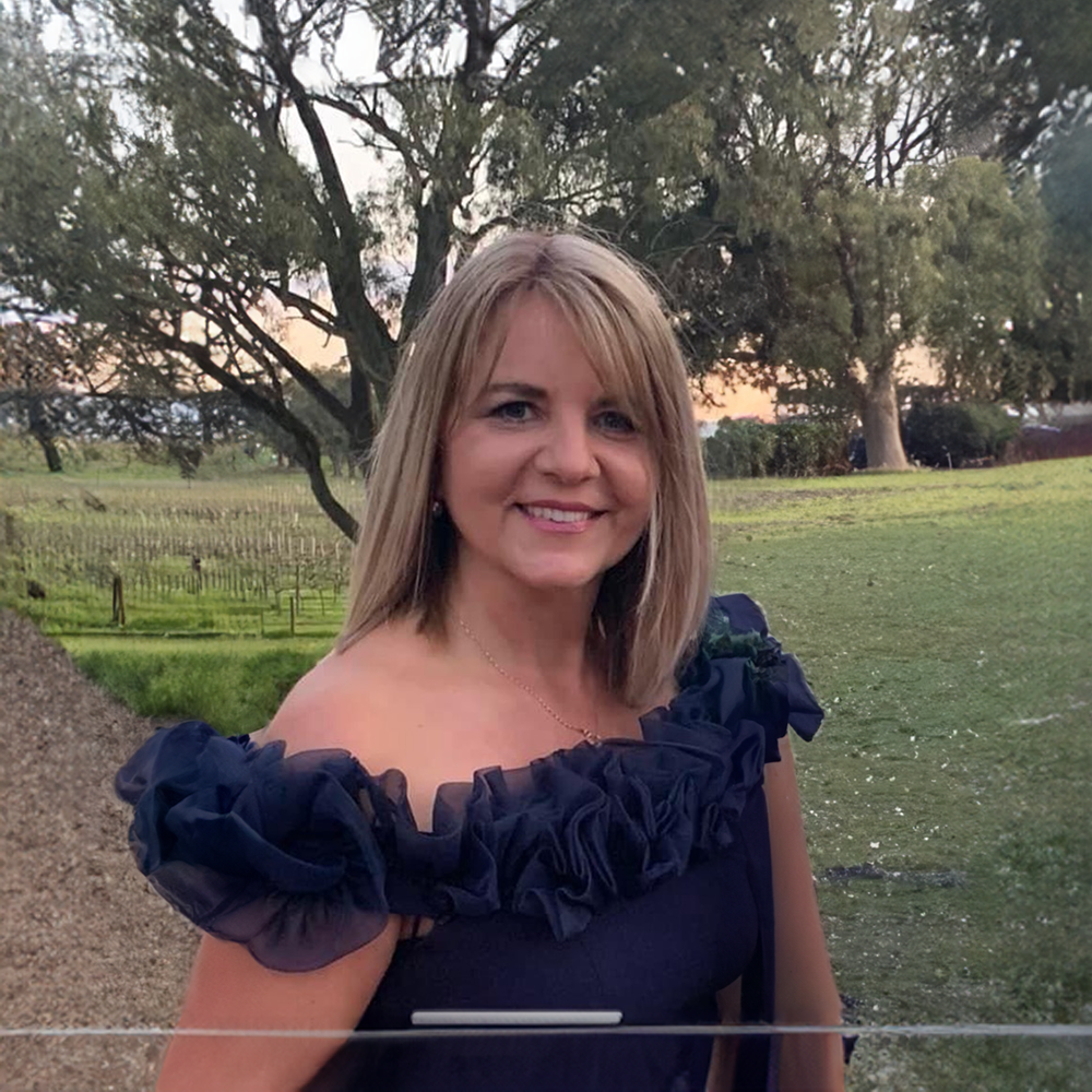 A woman with blonde hair smiling outdoors during sunset, wearing a navy blue dress with ruffled shoulder details.