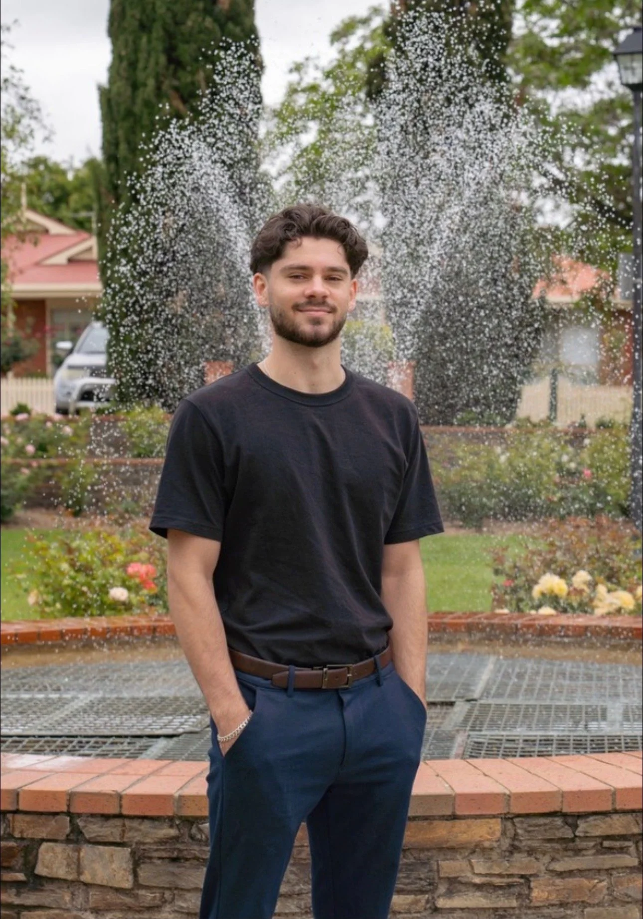 Young man standing in front of a fountain with water spraying behind him, in a park or garden setting with greenery and flowers.