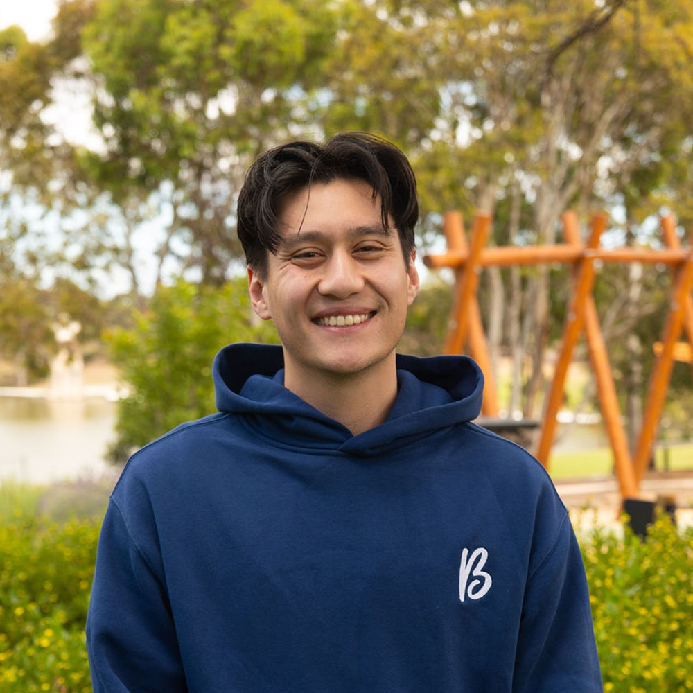 A young man with dark hair smiling, wearing a navy blue hoodie, standing outdoors in a park with trees, bushes, and a wooden structure in the background.