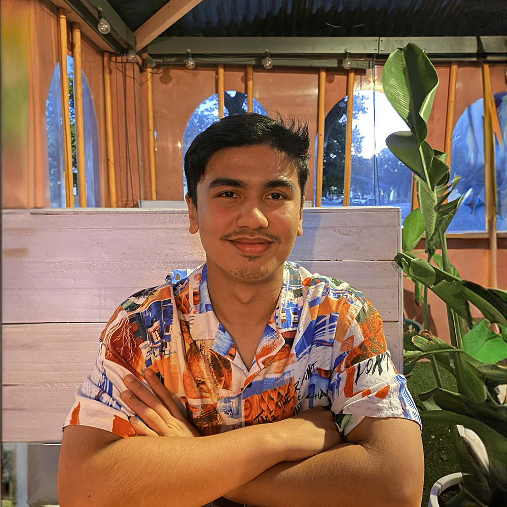 A young man with dark hair and a light beard, wearing a colorful short-sleeved button-up shirt, sits at a table with his arms crossed and is smiling at the camera. Behind him, there are large windows with an outdoor view of trees and evening sky, and a large green plant is visible on his right.