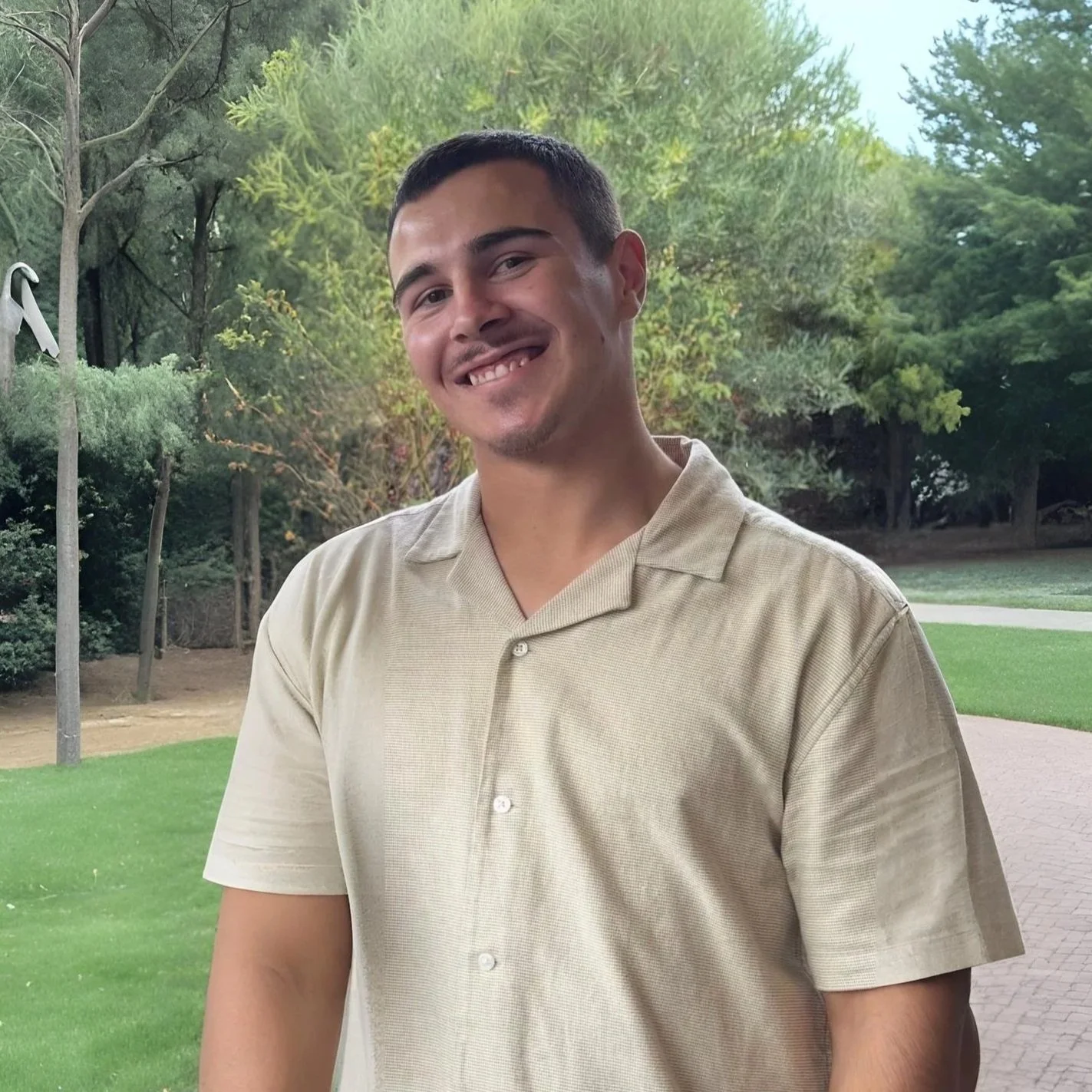 A young man with short dark hair, smiling, wearing a beige collared shirt, standing outdoors in a park with green trees and grass.