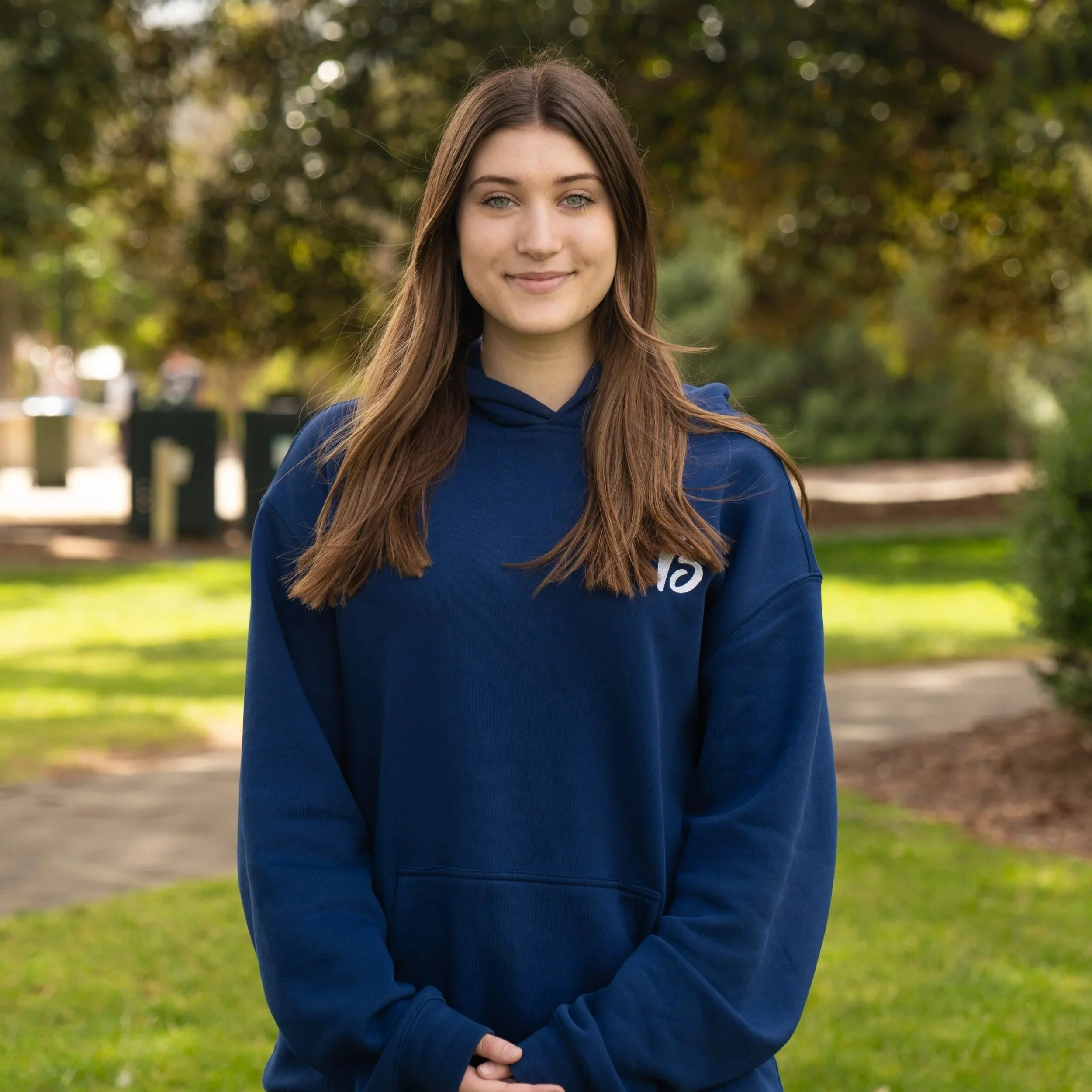 A young woman with light brown hair, styled in a side part, wearing a black jacket, smiling at the camera with a light blue background.