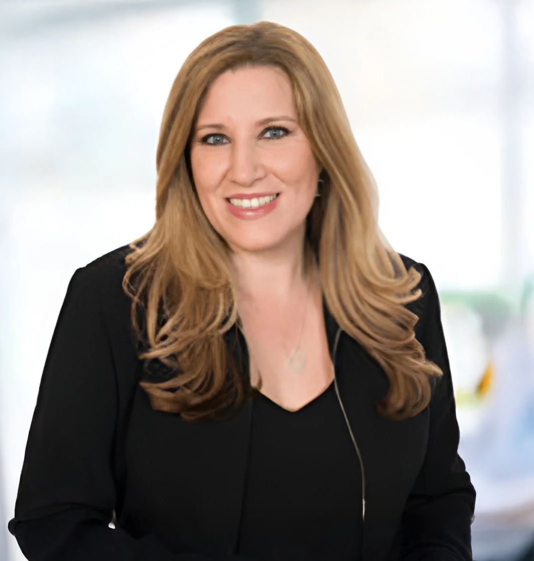 Portrait of a woman with long, wavy red hair, smiling, wearing a black top, with a blurred light-colored background.