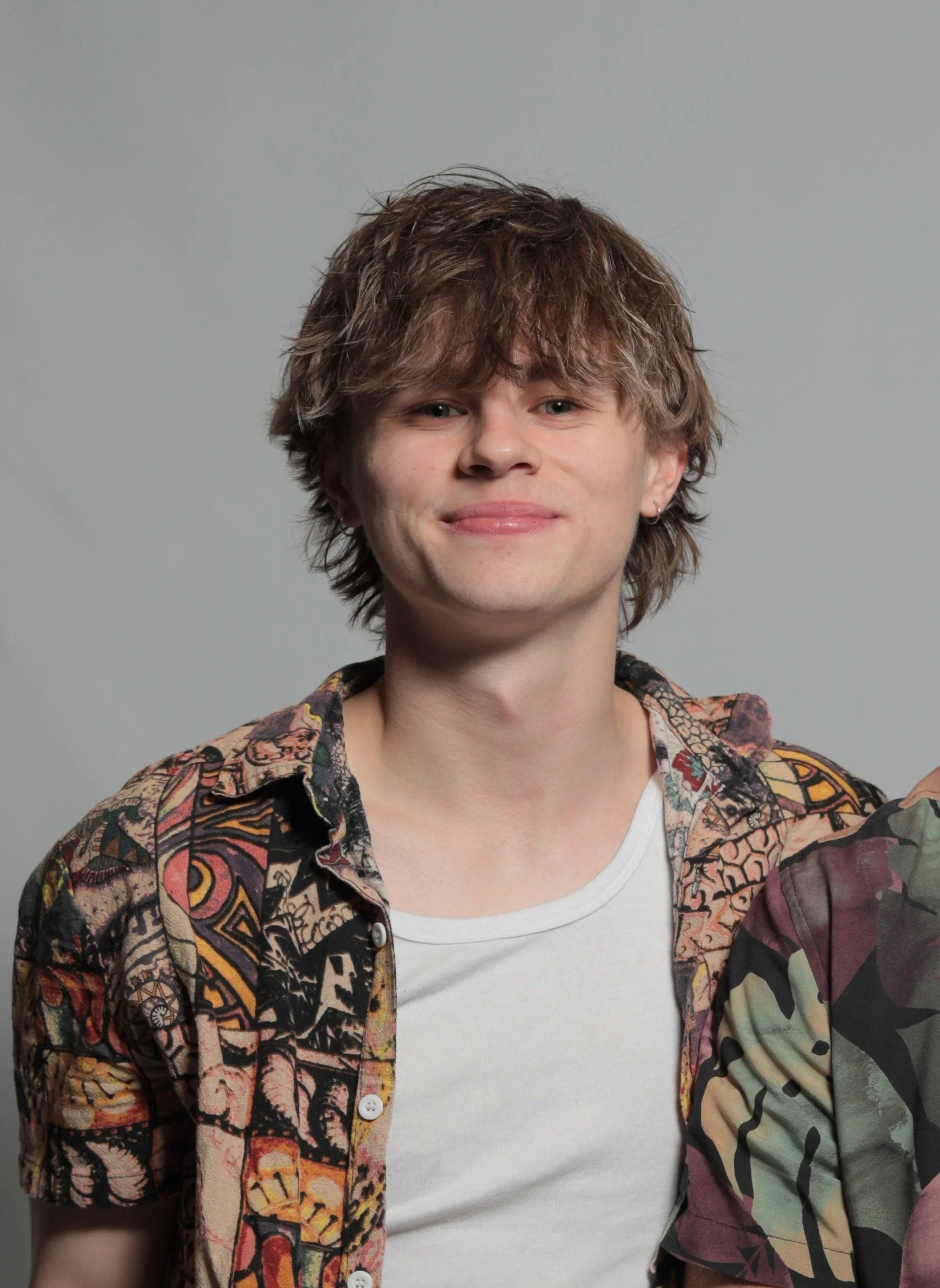 Young man with tousled brown hair wearing a colorful patterned shirt over a white t-shirt, smiling slightly against a plain gray background.