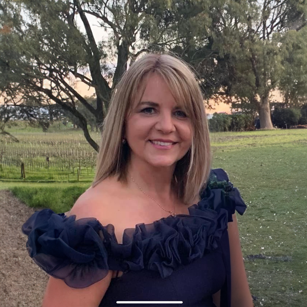 A woman with shoulder-length blonde hair, wearing a dark dress with ruffled sleeves, poses outdoors during sunset, with trees and a grassy landscape in the background.