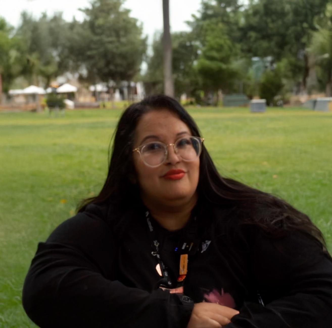 A woman with long dark hair, glasses, and red lipstick sitting outdoors on grass in a park, with trees and park structures in the background.