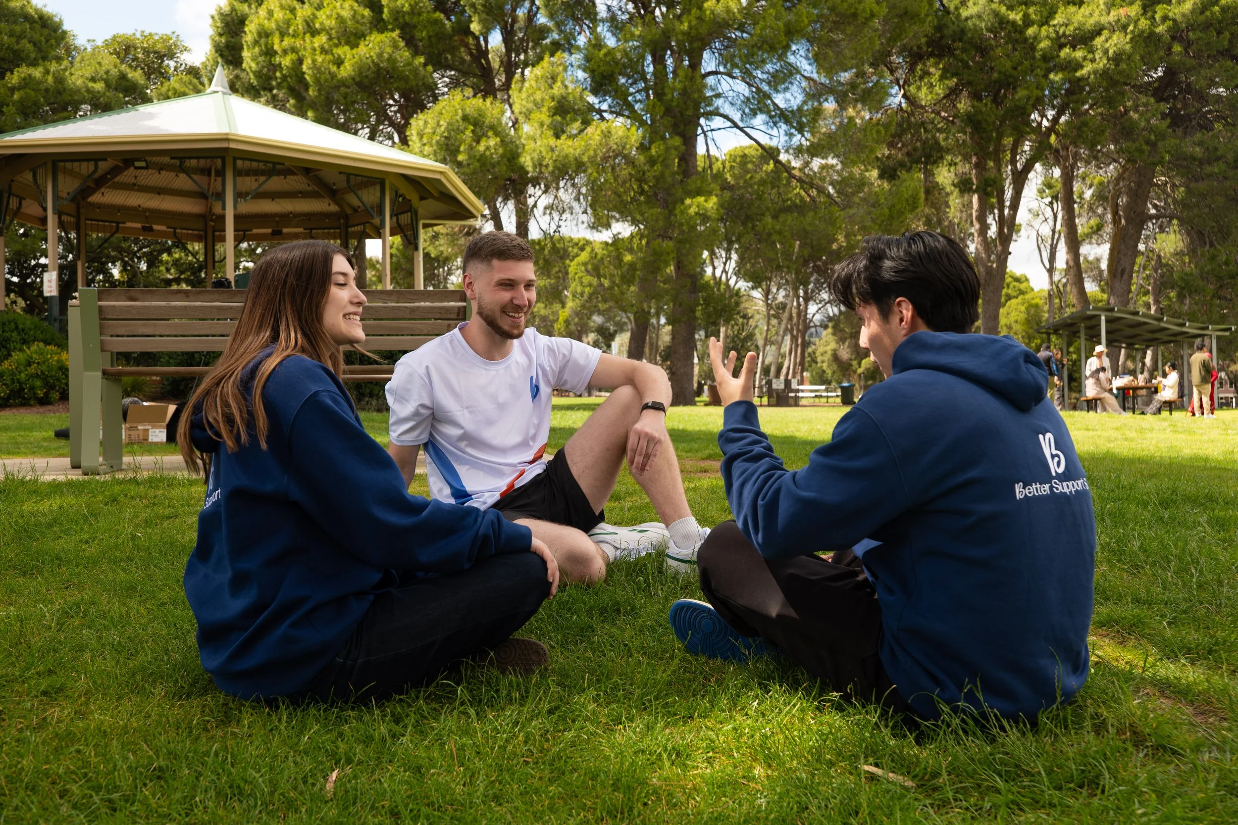 Three young adults sitting on the grass in a park, engaging in a lively conversation, with trees and park structures in the background.