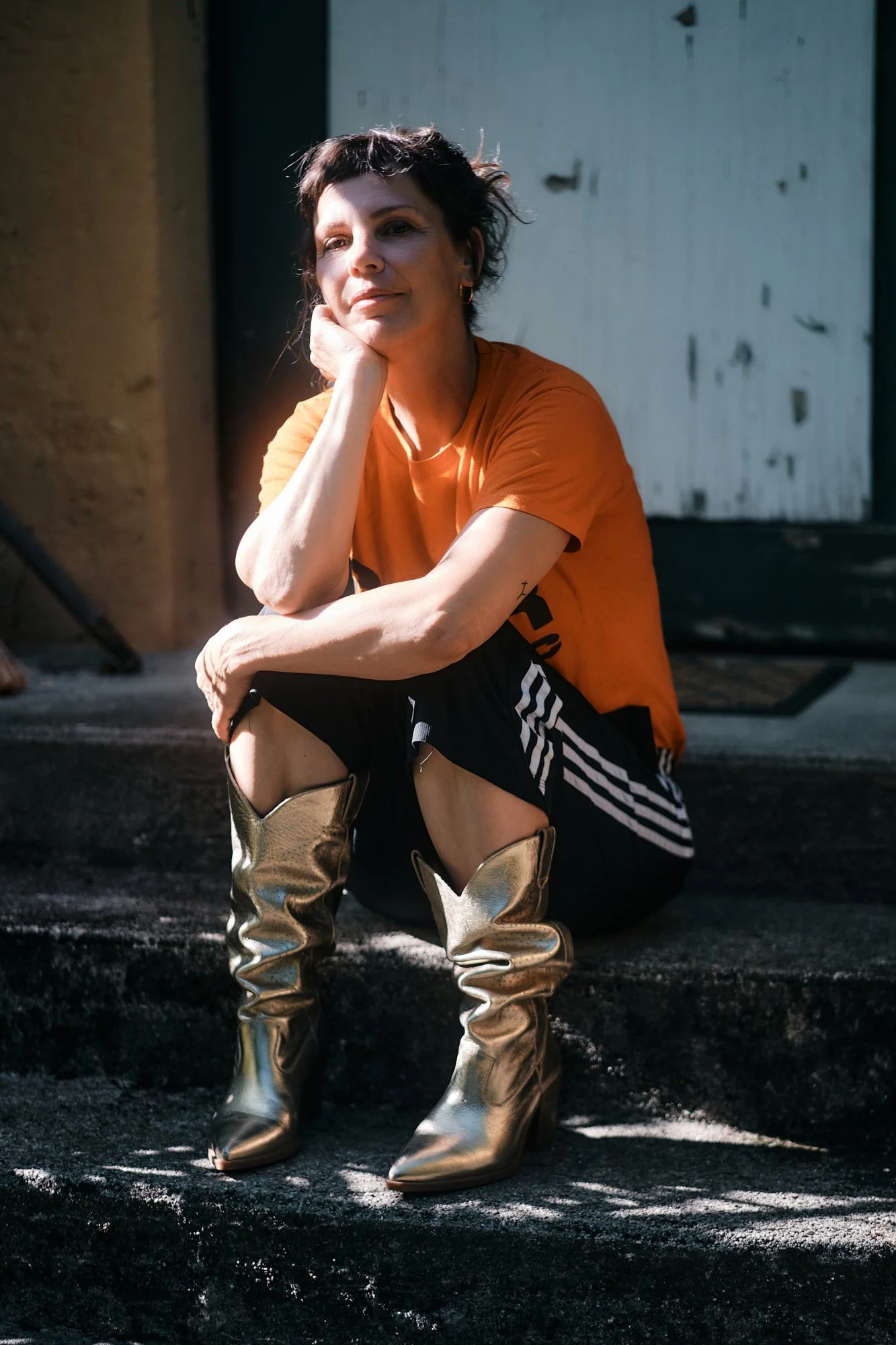 Frau sitzt auf einer schwarzen Treppe und trägt eine orange T-Shirt, schwarze Shorts mit weißen Streifen und goldene Stiefel, blickt nach oben.