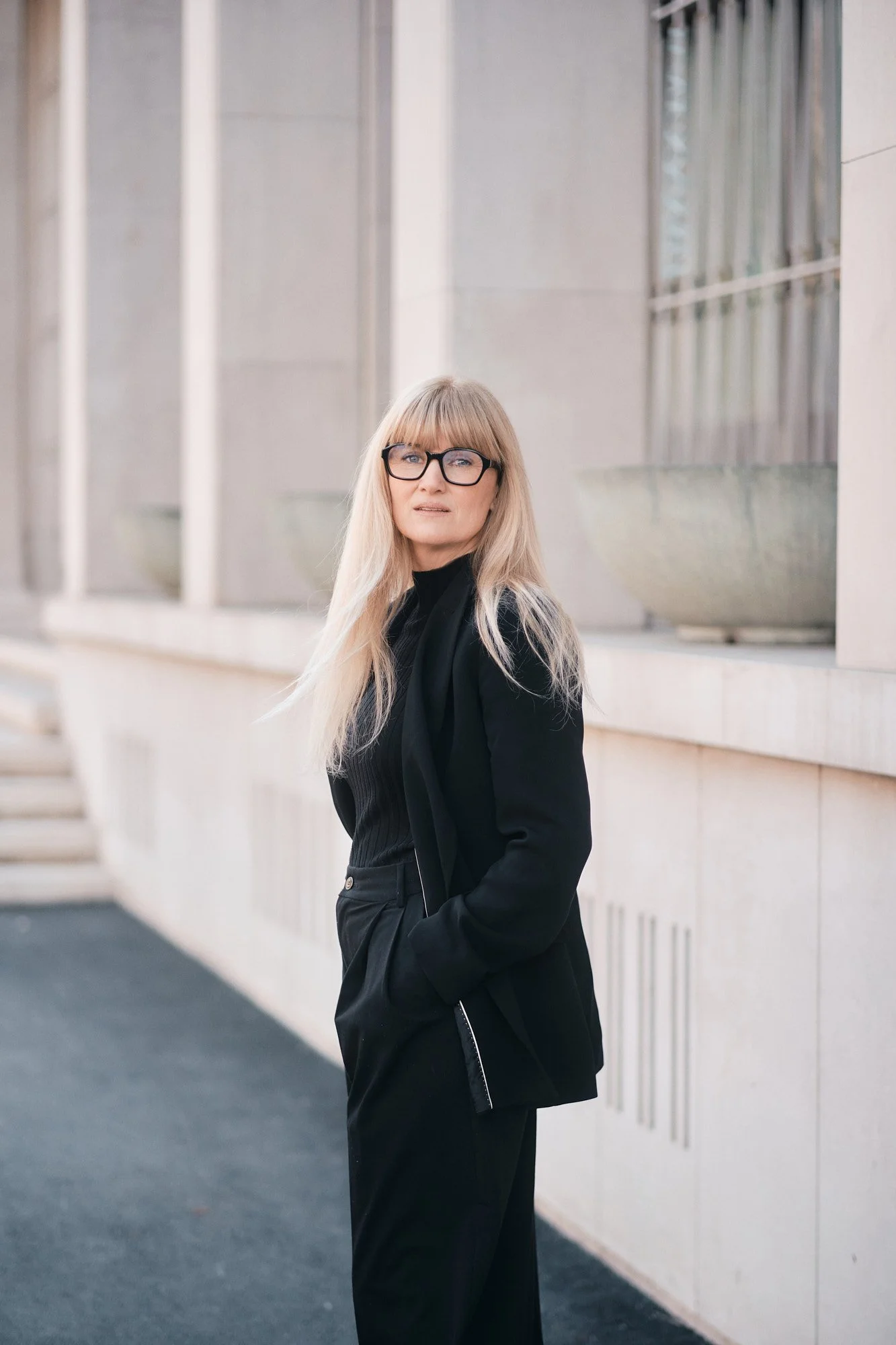 Eine blonde Frau mit Brille in einer schwarzen Jacke und schwarzen Hose steht vor einem modernen Gebäude mit Steinfassade.