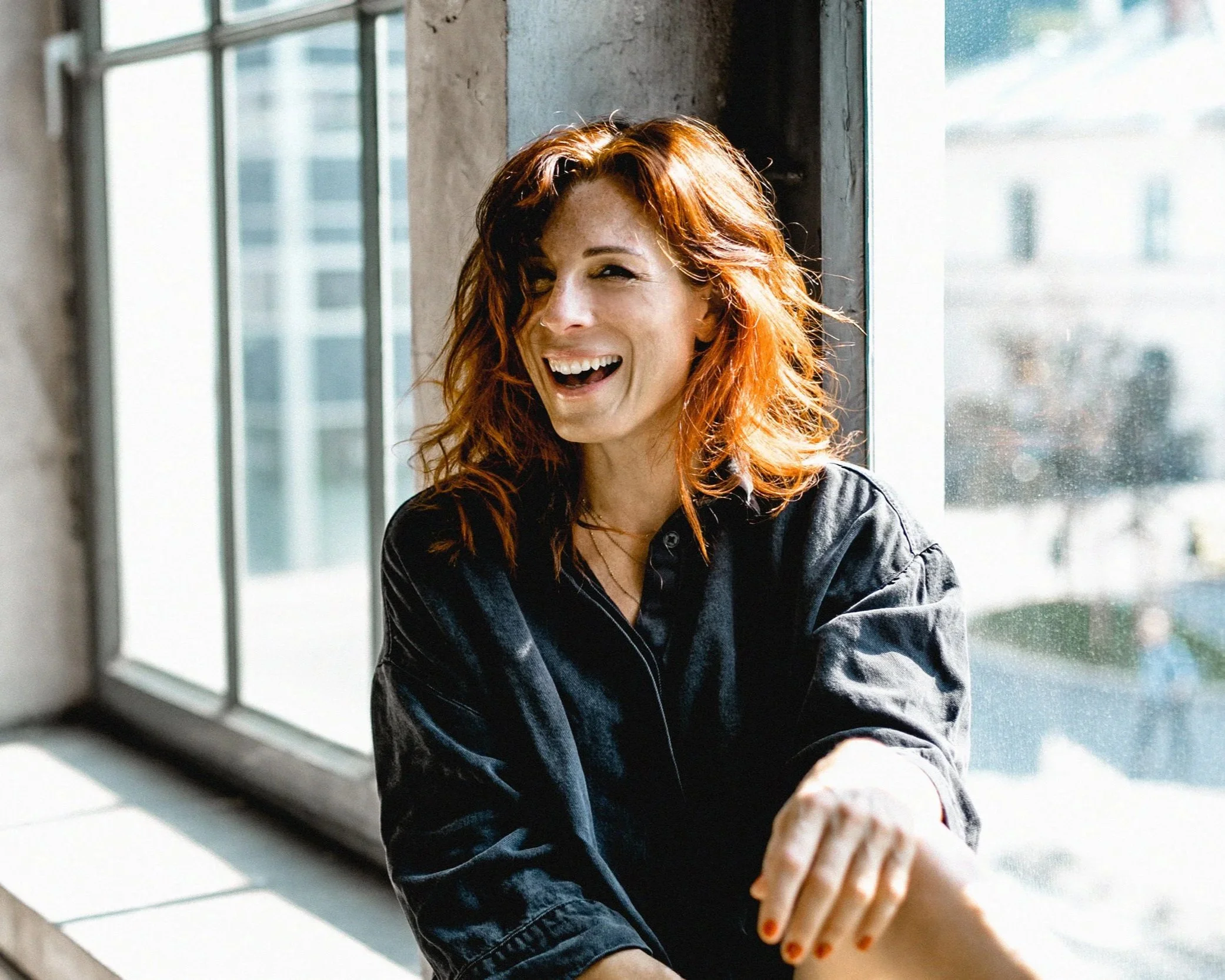 Eine lachende Frau mit roten Haaren sitzt an einem Fenster in einem hellen, modernen Raum.