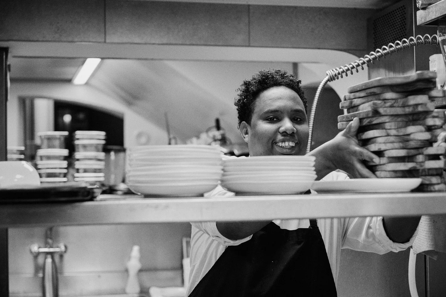 Ein Lächelnder Koch in einer Küche, der eine große Stapel Pizzastücke trägt, mit mehreren Schüsseln und anderen Küchenutensilien im Hintergrund.