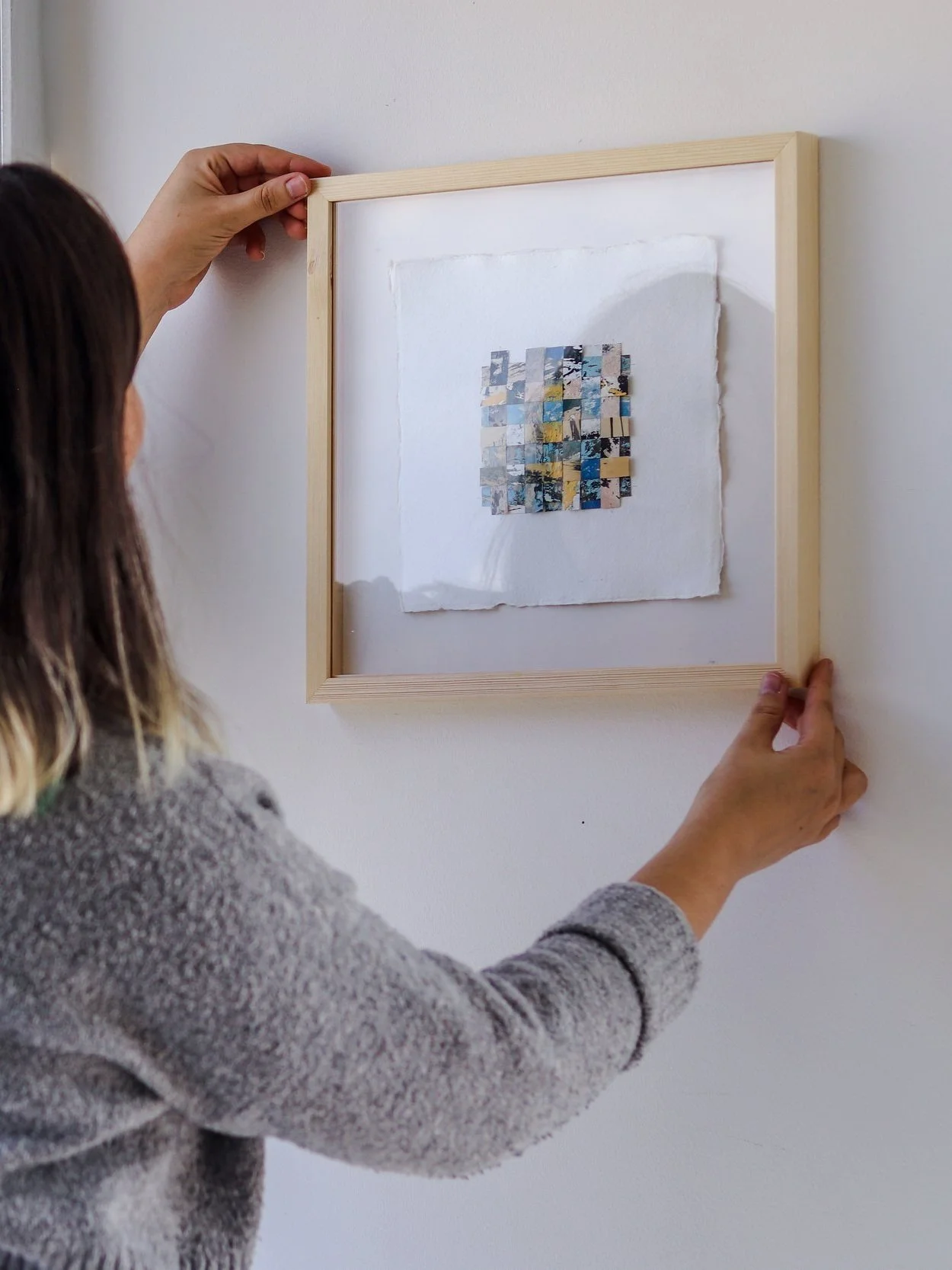 Artist installing framed Woven Impressions: Woven monotype artwork, with textured oil paint paper strips sewn in an interlaced pattern featuring yellow, light blue, dark blue, pink, grey, and white.