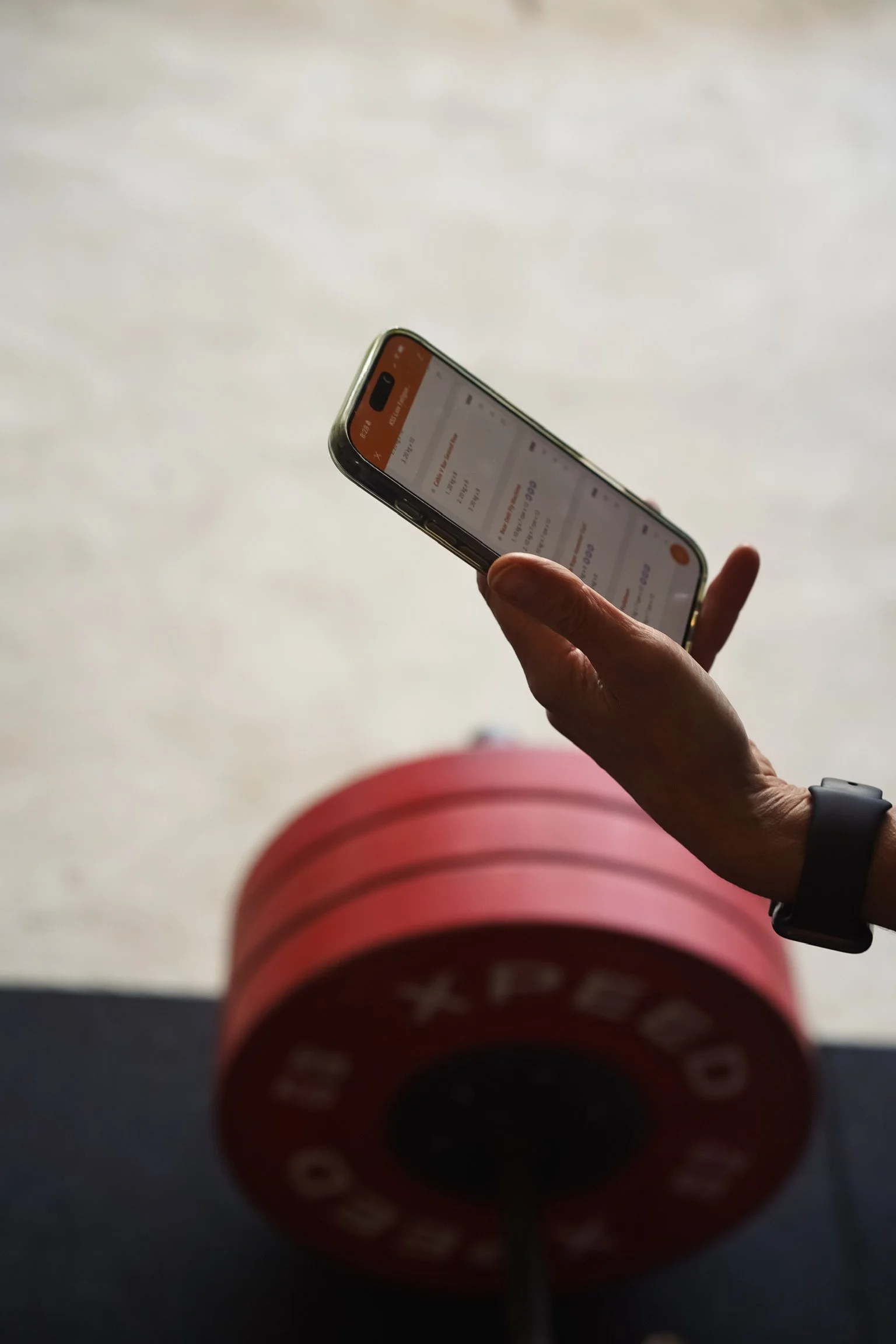 Person holding a smartphone over their knee, with red and black workout equipment on the floor and a light-colored wall in the background.