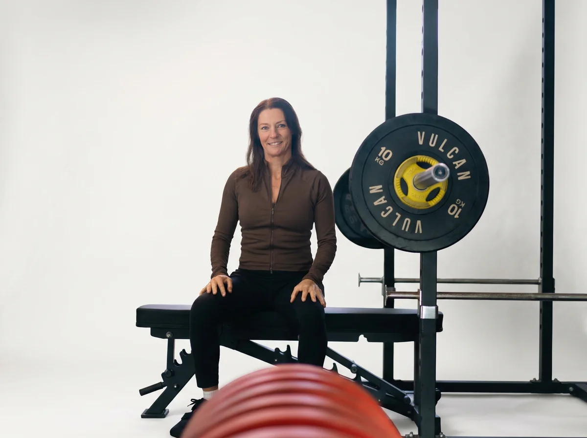 A woman sitting on a workout bench in a gym, with weights and a squat rack nearby.