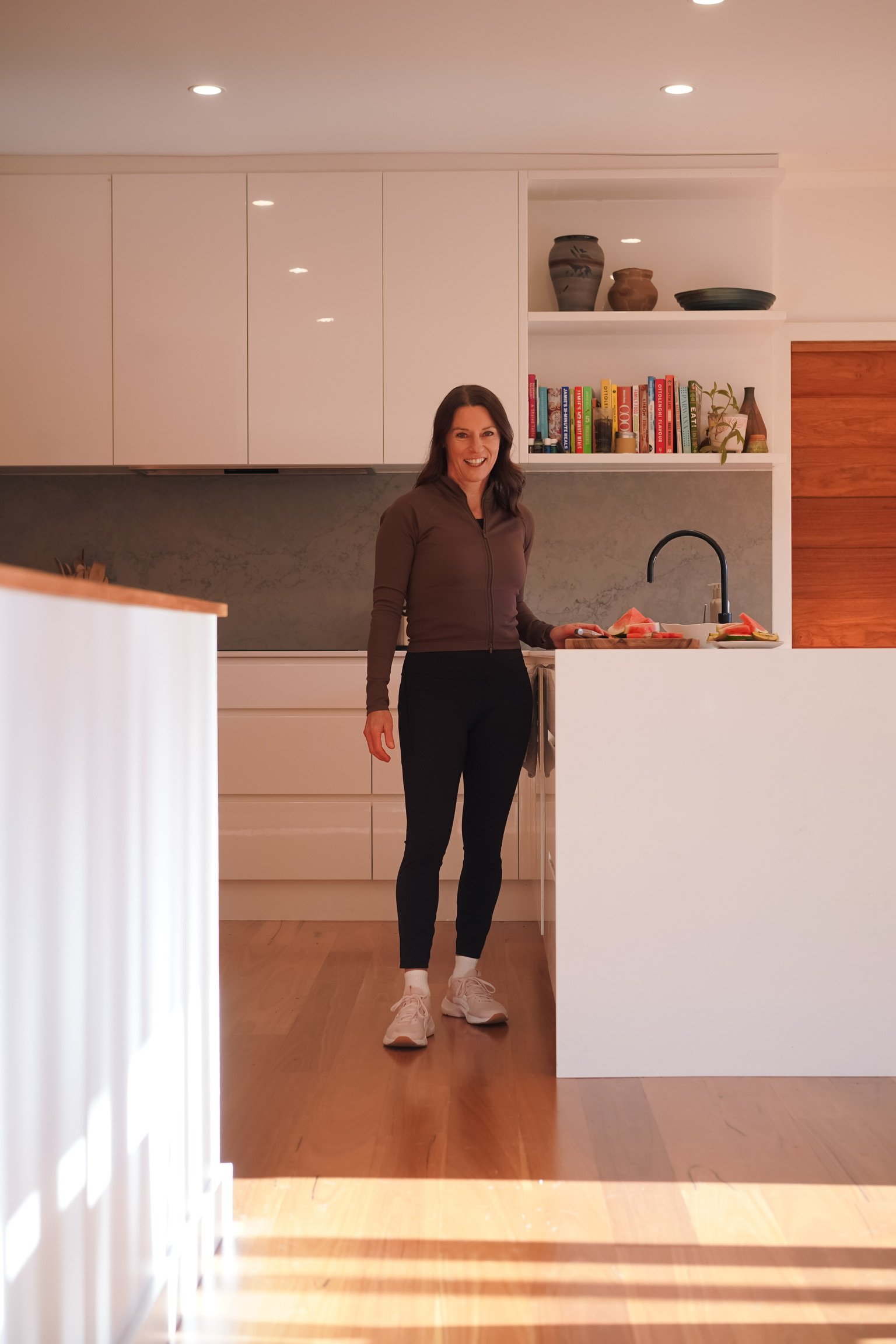 A woman standing in a modern kitchen with white cabinets, wooden floors, a marble backsplash, and a small open shelf with vases and books. She is smiling, holding a plate of sliced watermelon, wearing a brown zip-up hoodie, black pants, white sneakers, and white socks.