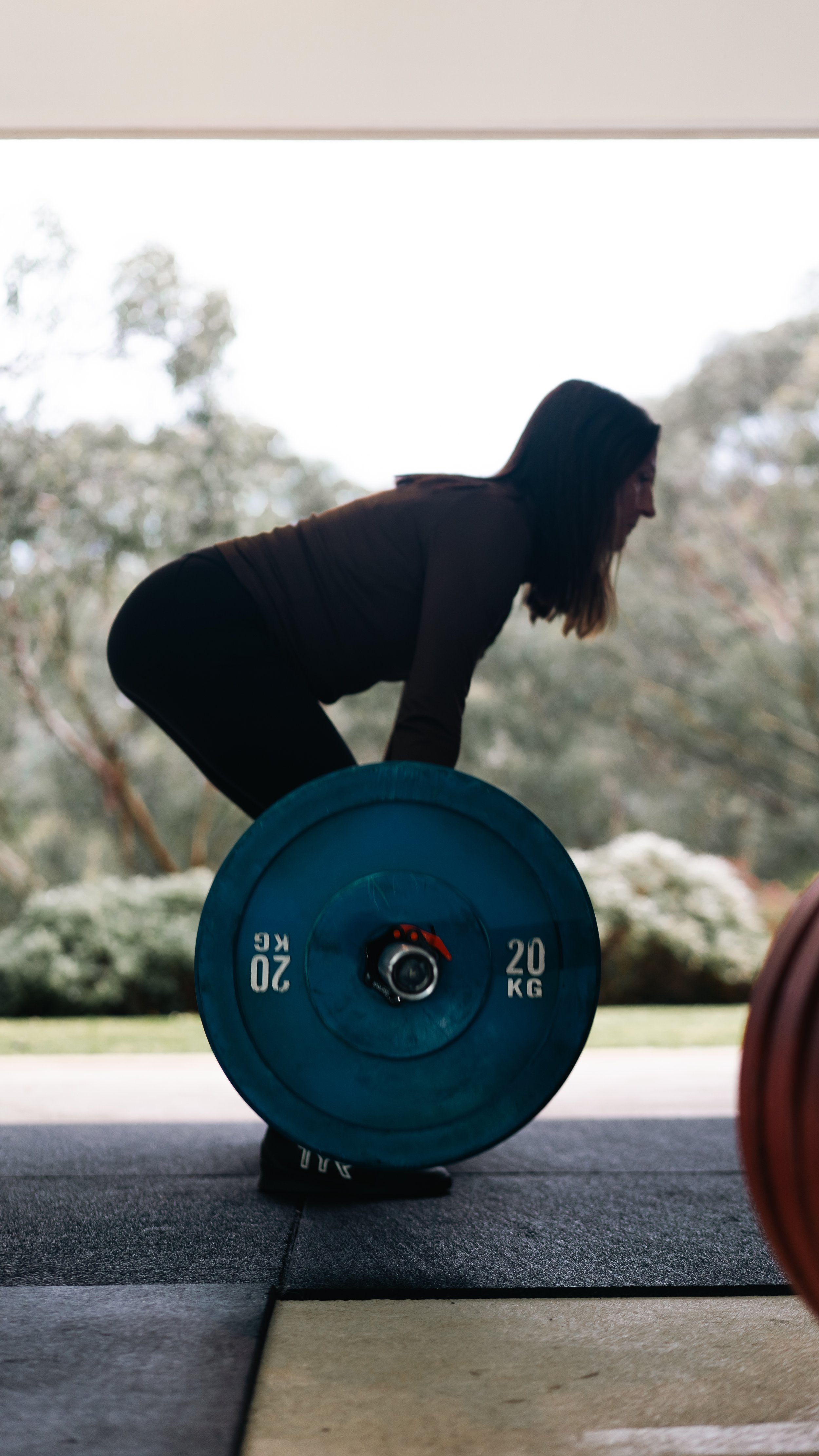 A woman in a black long-sleeve shirt and black pants lifting a 20 kg blue weight plate during exercise in a gym with large windows and outdoor trees in the background.